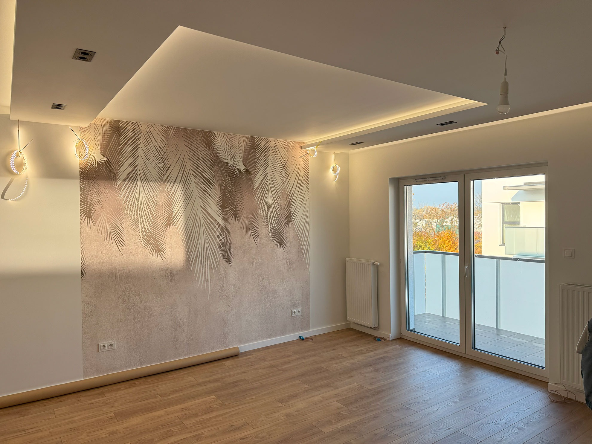 Jasne wnętrze po remoncie: parkiet, ściana z motywem liści palmowych, sufit podwieszany z oświetleniem, okno balkonowe z widokiem na zewnątrz. Częściowo zamontowane lampy.