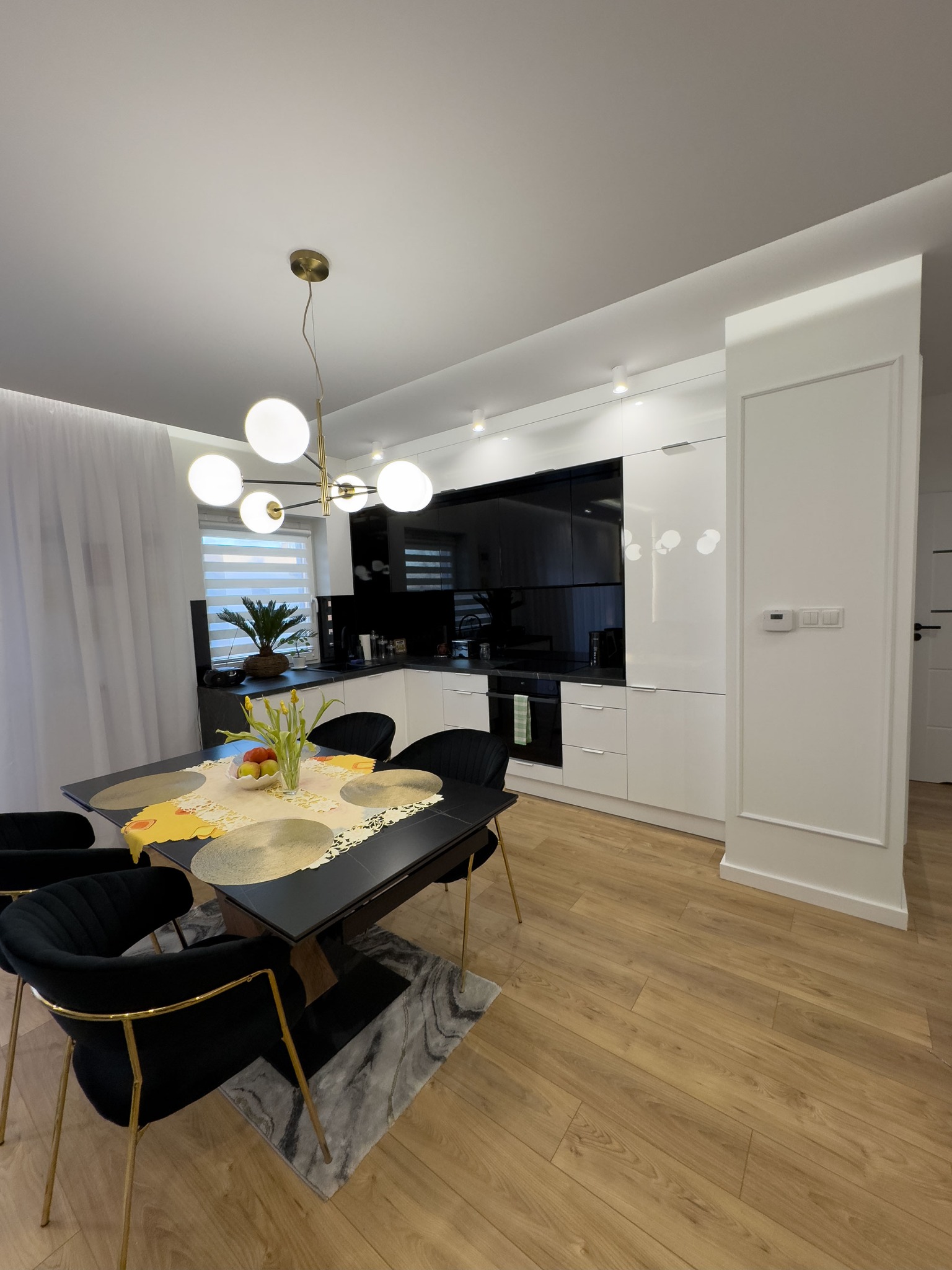 Nowoczesna kuchnia z czarno-białymi szafkami i blatem, oświetlona kulistą lampą. Stół z czarnymi krzesłami na złotych nogach i kwiatami. Parkiet na podłodze.