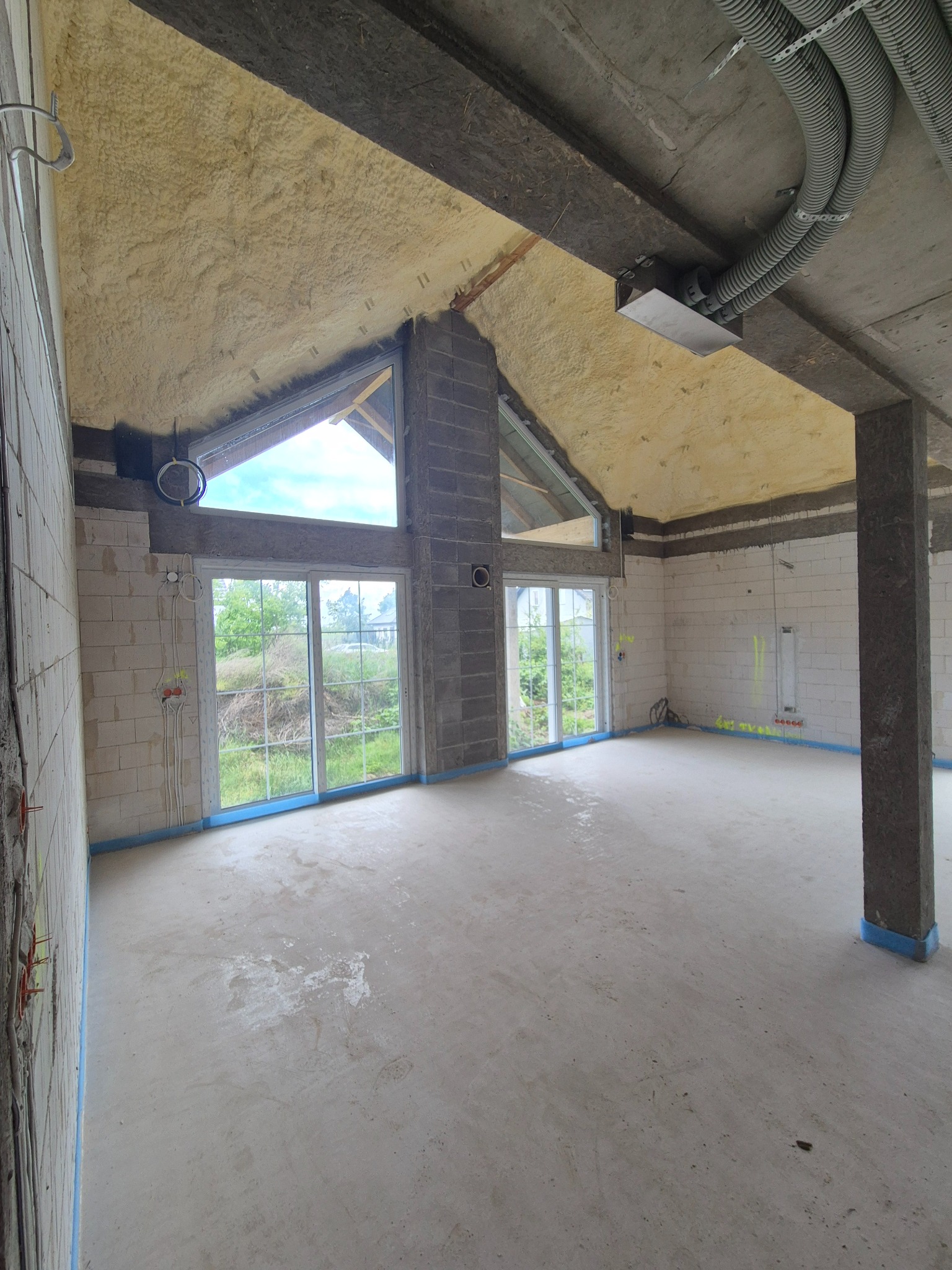 Wnętrze domu w trakcie ocieplania poddasza pianą PUR. Widoczne okna dachowe, ściany z bloczków i instalacje elektryczne. Posadzka wylewana, przygotowana do dalszych prac.