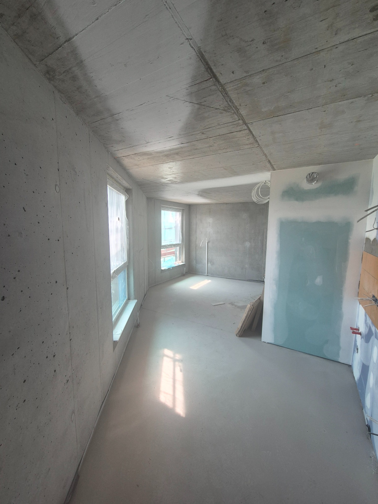 Surowe wnętrze w trakcie prac wykończeniowych: betonowe ściany, okna zabezpieczone folią, częściowo położona gładź na ścianie, widoczne instalacje elektryczne.