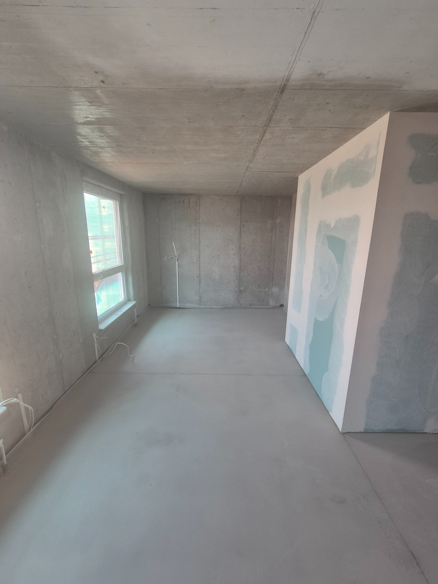 Surowe wnętrze w trakcie wykańczania: betonowe ściany i podłoga, okno z folią ochronną, płyta gipsowa z widocznymi śladami szpachlowania. Minimalistyczny, industrialny styl.