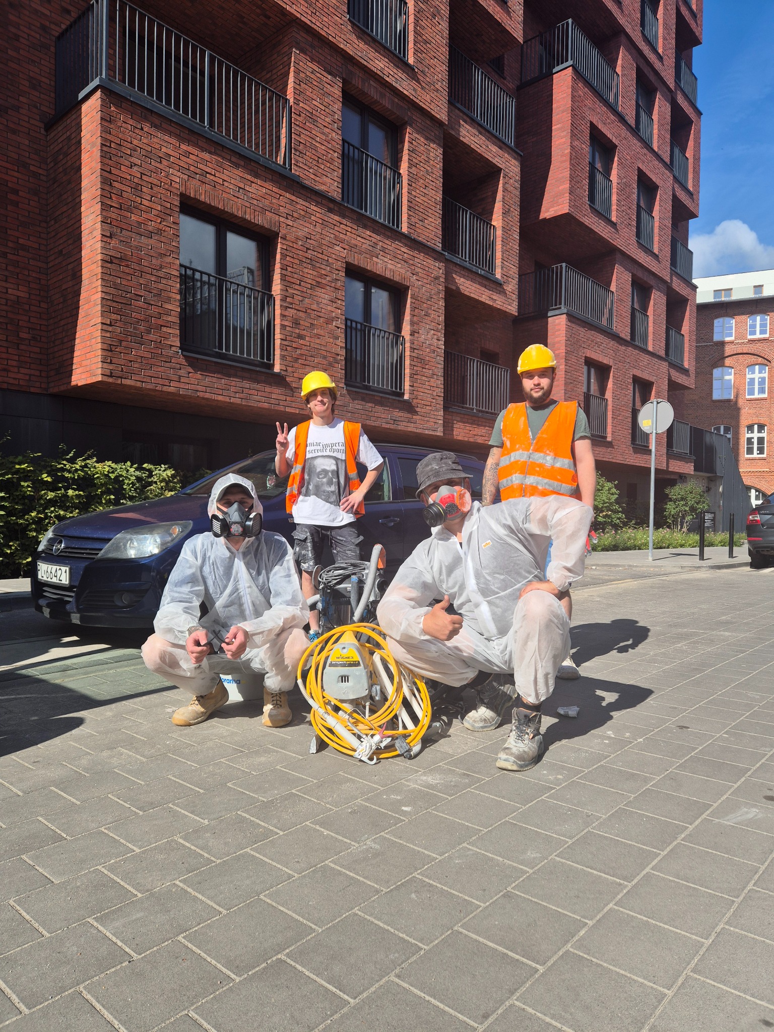 Ekipa w kombinezonach i kaskach pozuje z maszyną natryskową na tle budynku z czerwonej cegły. Widoczny samochód i fragment chodnika.