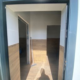 PrishTech-Bud Vladyslav Pryshchepa - Wnętrze łazienki z wykończonymi płytkami imitującymi drewno na ścianach i podłodze, widoczne przez otwarte, grafitowe drzwi. W tle widoczna miotła.