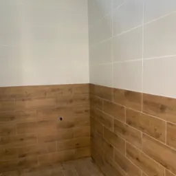 Wykończona łazienka z płytkami imitującymi drewno na dole i jasnymi płytkami na górze ścian. Widoczny fragment sufitu z płytą karton gips.