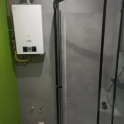 Szary prysznic z czarną ramą i biały piec gazowy Ferroli na zielonej ścianie. Widoczne rury i płytki. Minimalistyczne wykończenie łazienki.