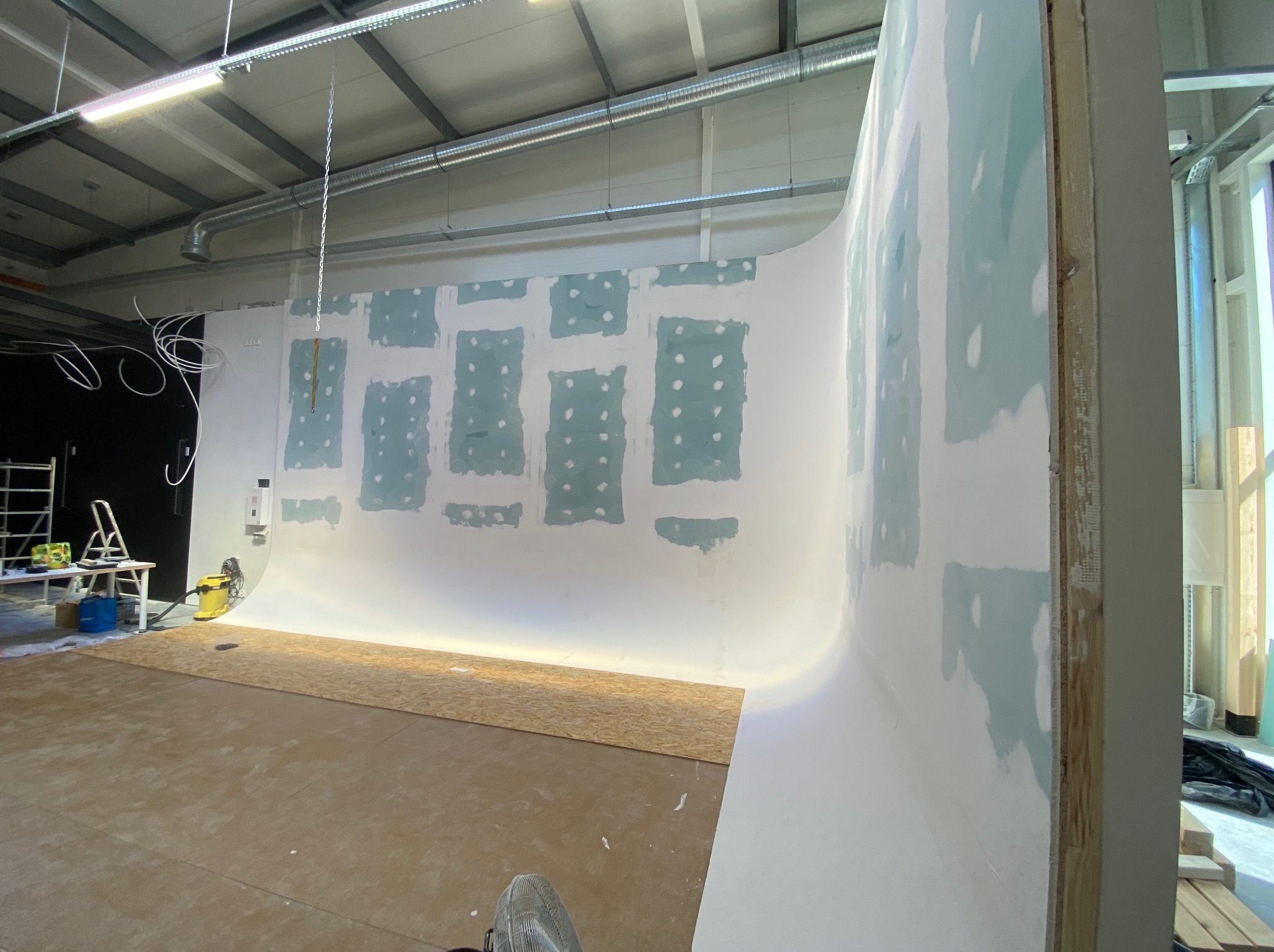 Wnętrze w trakcie prac: ściana z płyty karton-gips z widocznymi miejscami szpachlowania, podłoga częściowo wyłożona płytą OSB, widoczne narzędzia i instalacje.