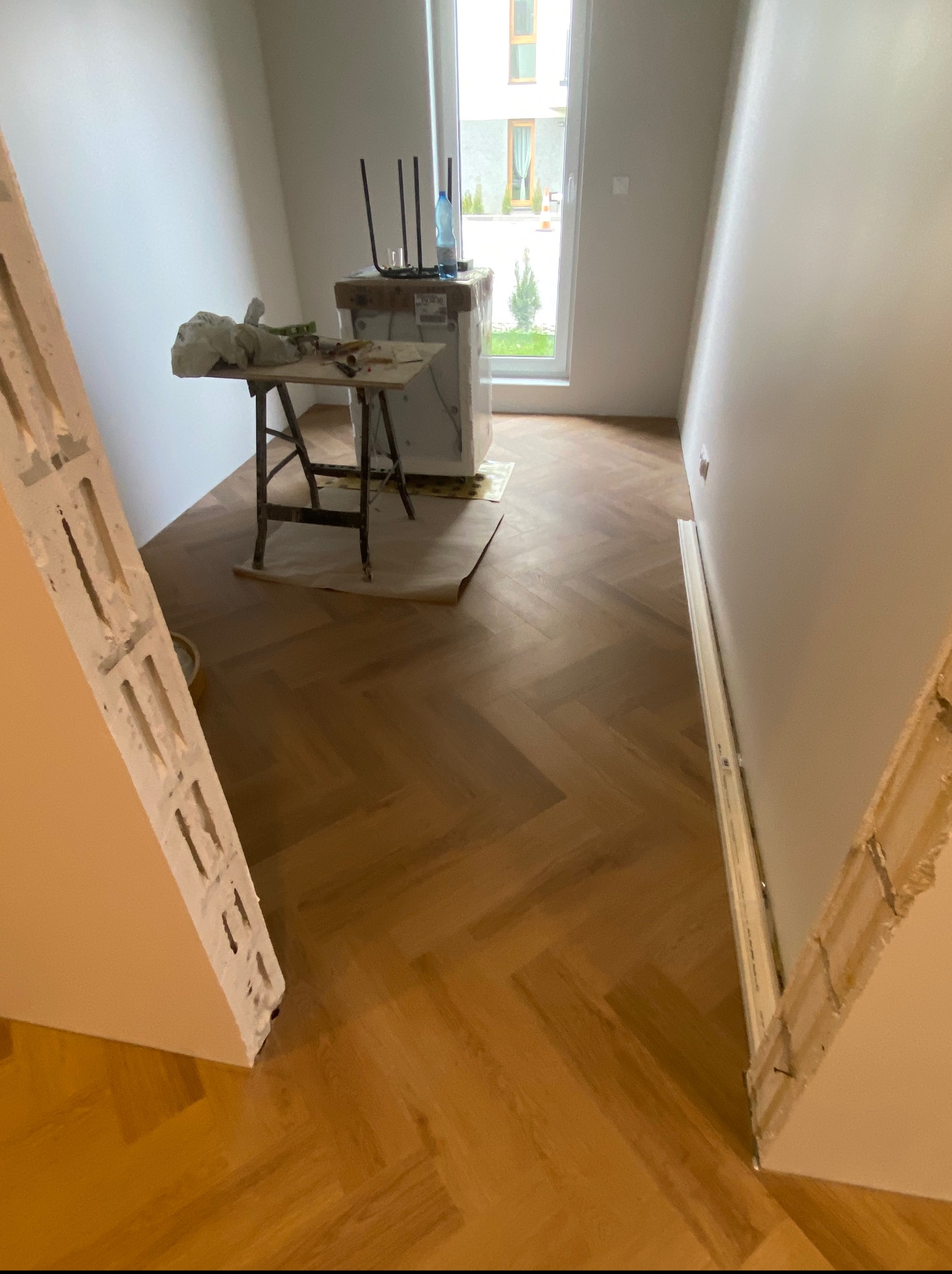Ułożony parkiet w jodełkę w trakcie wykańczania. W tle widoczne narzędzia pracy na stole. Fragment ściany z niedokończonym wykończeniem. Jasne wnętrze z oknem.