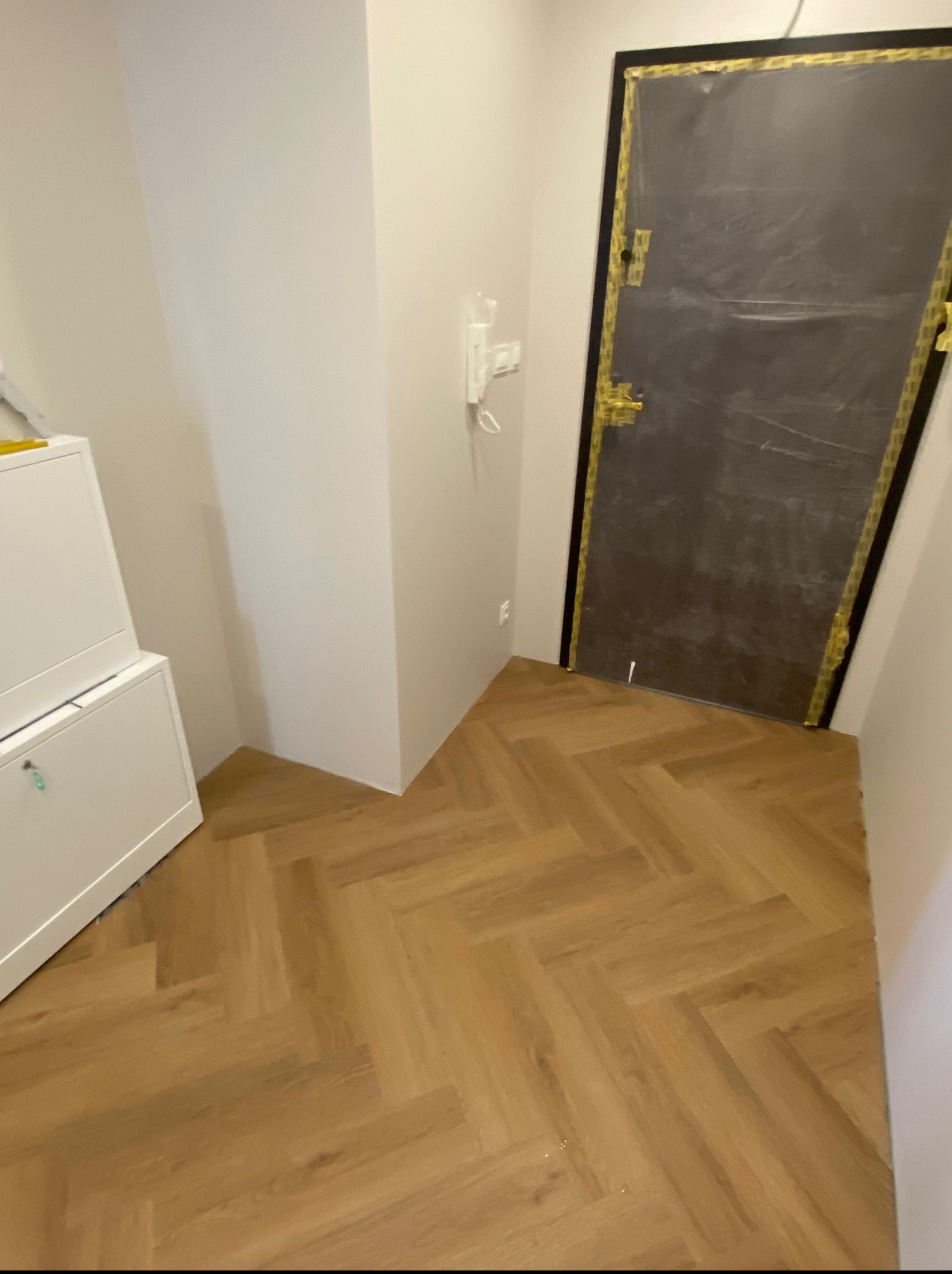 Ułożona podłoga z paneli w jodełkę w jasnym korytarzu, drzwi zabezpieczone folią i taśmą malarską, biała szafka w rogu. Widok na gotową posadzkę.