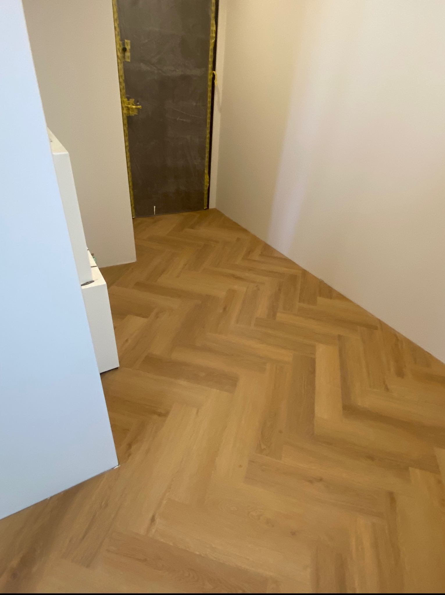 Ułożone panele podłogowe w jodełkę w jasnym odcieniu drewna, widok na fragment ściany i zabezpieczone taśmą drzwi w trakcie prac wykończeniowych.