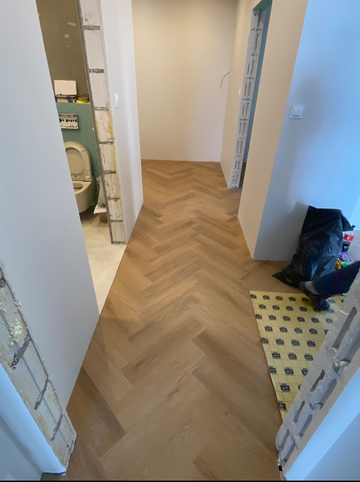 Ułożony parkiet w jodełkę w korytarzu w trakcie remontu. Widoczne materiały budowlane i przygotowane podłoże pod panele. Częściowo wykończone ściany.