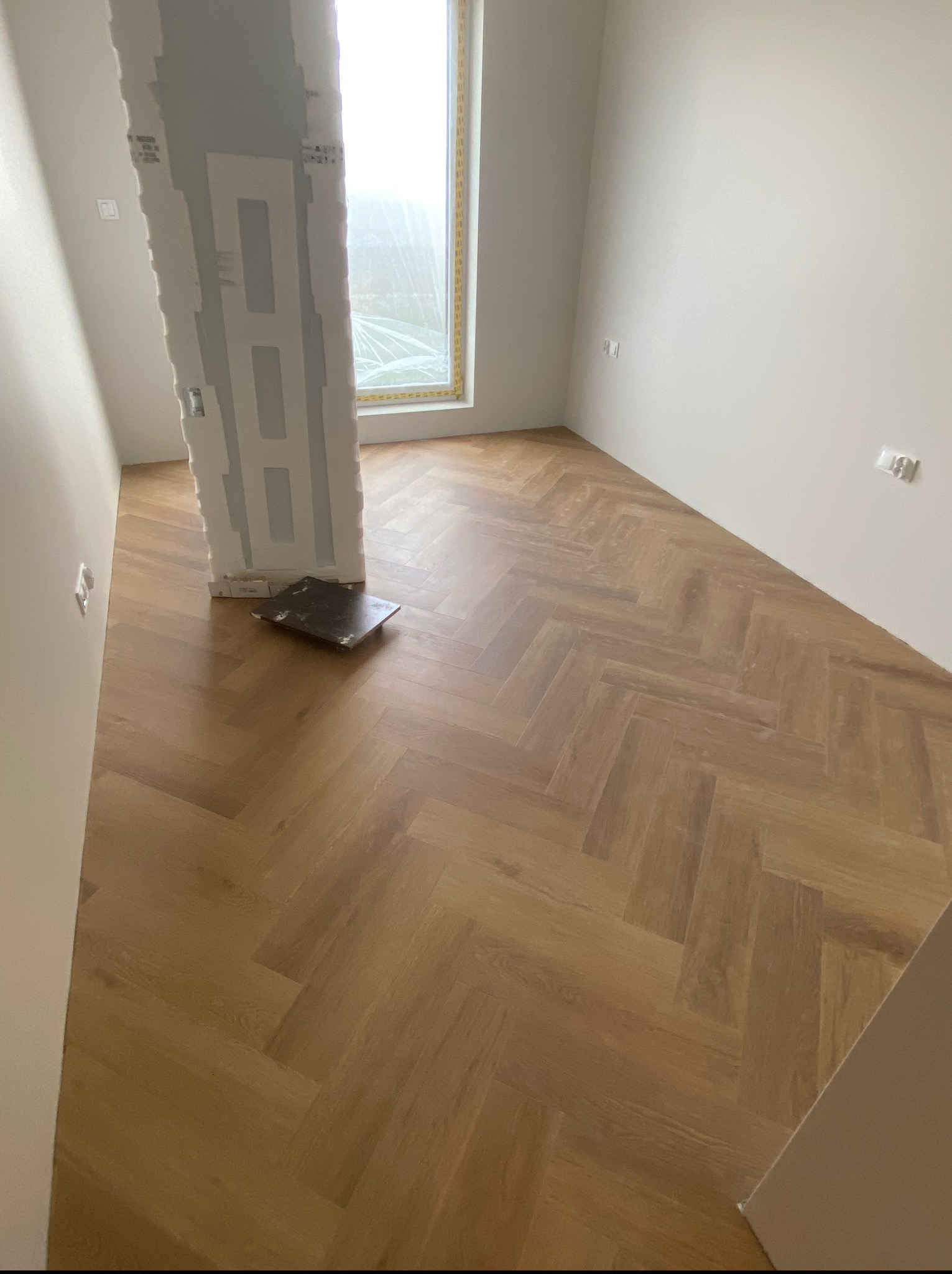 Parkiet ułożony w jodełkę francuską w jasnym, nowoczesnym wnętrzu z płytą gipsową przy ościeżnicy. Widoczne gniazdka elektryczne.