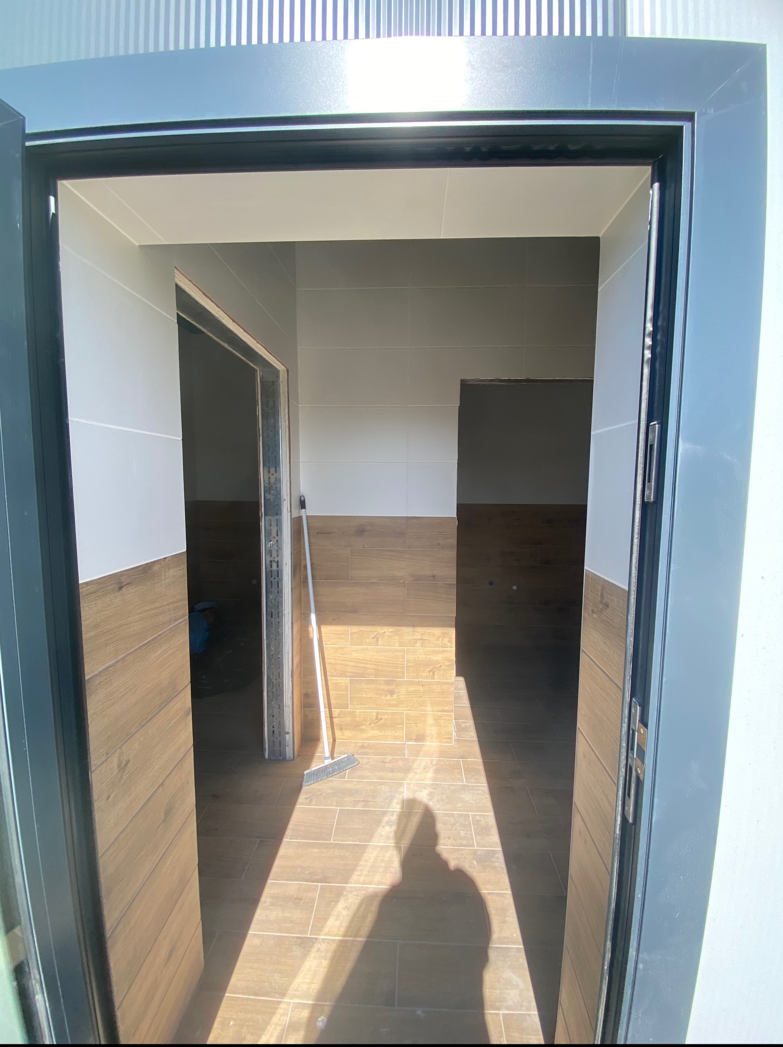 Wnętrze łazienki z wykończonymi płytkami imitującymi drewno na ścianach i podłodze, widoczne przez otwarte, grafitowe drzwi. W tle widoczna miotła.