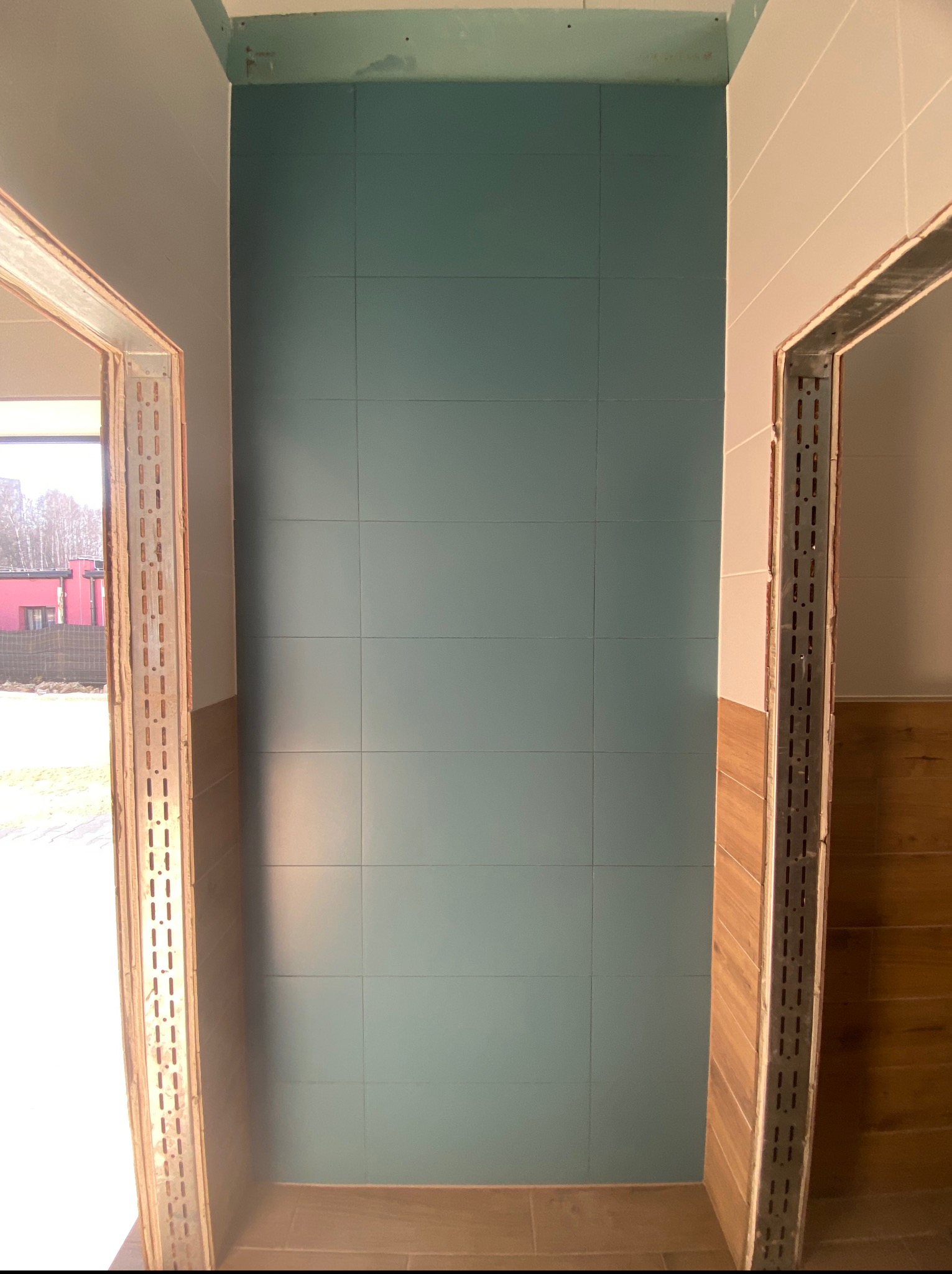 Ściana wyłożona turkusowymi płytkami w trakcie wykańczania łazienki. Widoczne metalowe profile ościeżnic i fragment drewnopodobnych płytek na ścianie bocznej.