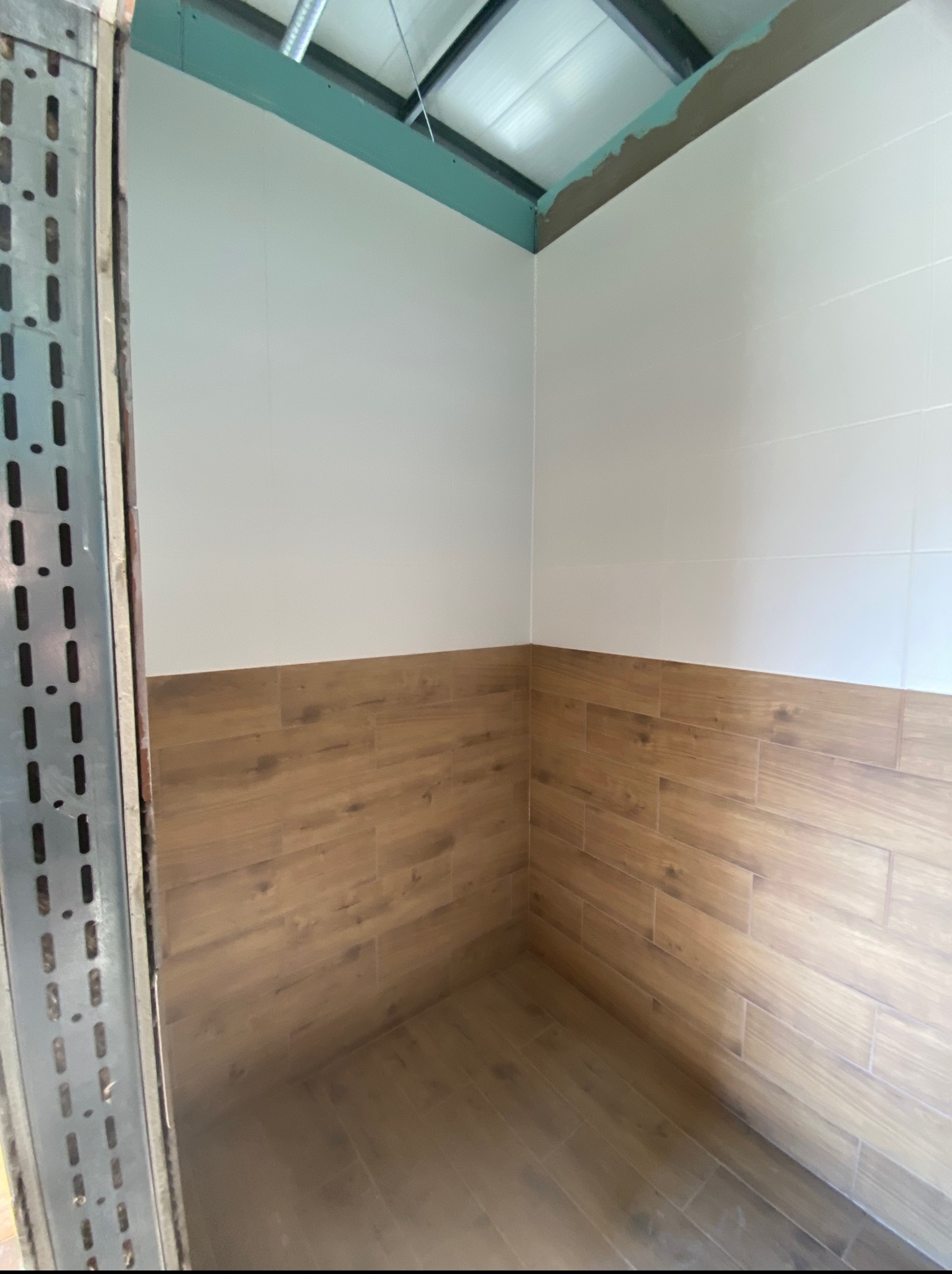 Wykończona łazienka z białymi i drewnopodobnymi płytkami, widok na narożnik i fragment metalowej konstrukcji. Świeżo położone płytki, widoczne łączenia.