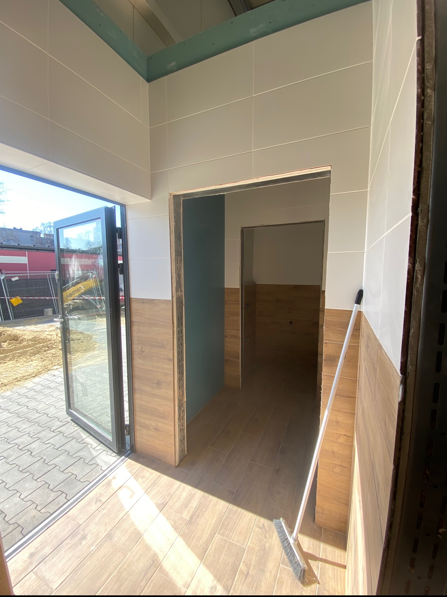 Wnętrze w trakcie prac wykończeniowych: jasne płytki na ścianach, drewnopodobne panele na podłodze. Widoczny otwarty szklany portal i szczotka oparta o ścianę.