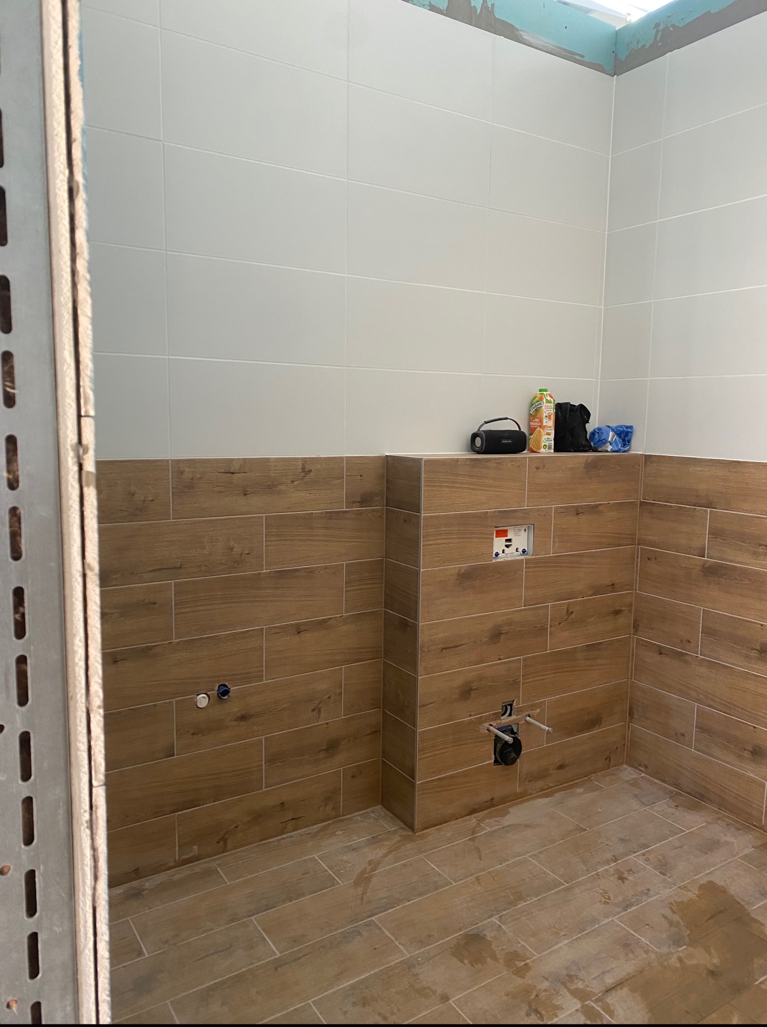 Łazienka w trakcie wykańczania: białe i drewnopodobne płytki, widoczne instalacje wodne. Na górze leżą akcesoria, a w rogu metalowy profil.