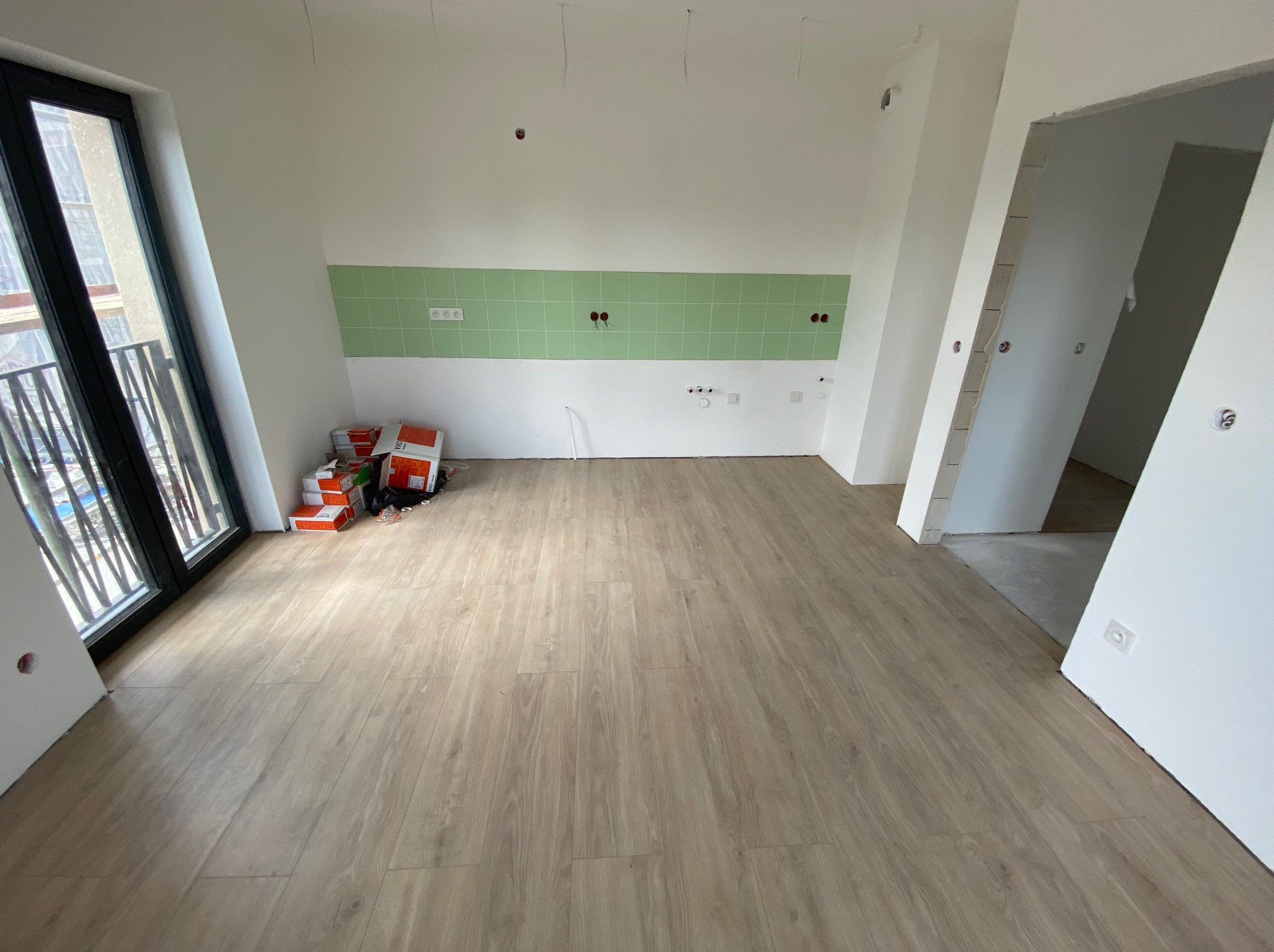 Nowe panele podłogowe w jasnym odcieniu drewna w wykończonym wnętrzu z zielonymi płytkami. Widoczne puszki elektryczne i okno z balkonem.