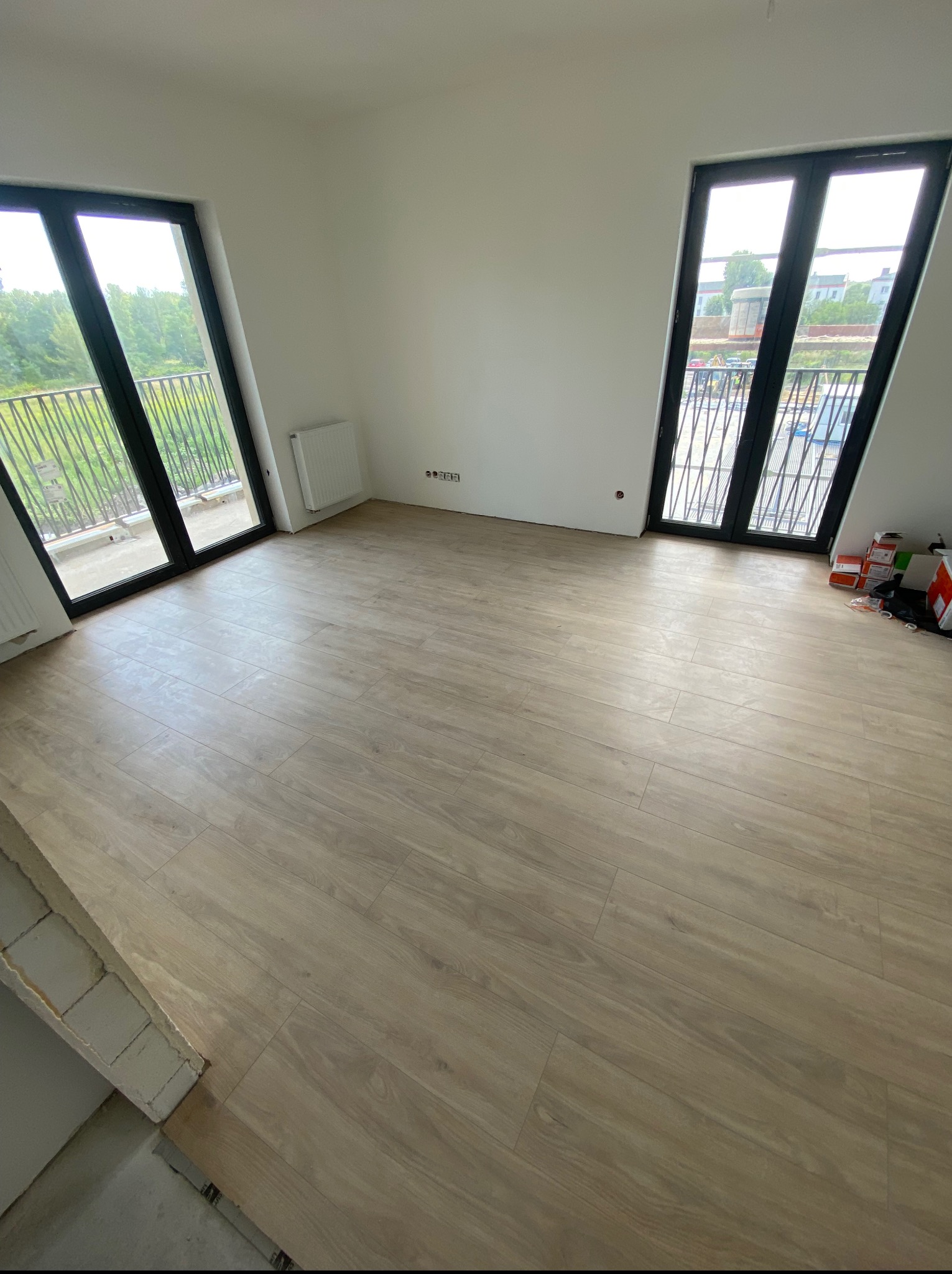 Jasne panele podłogowe ułożone w pustym pokoju z dwoma balkonami. Widoczne skrzynki na podłodze. Świeżo wykończone wnętrze z grzejnikiem i gniazdkami.