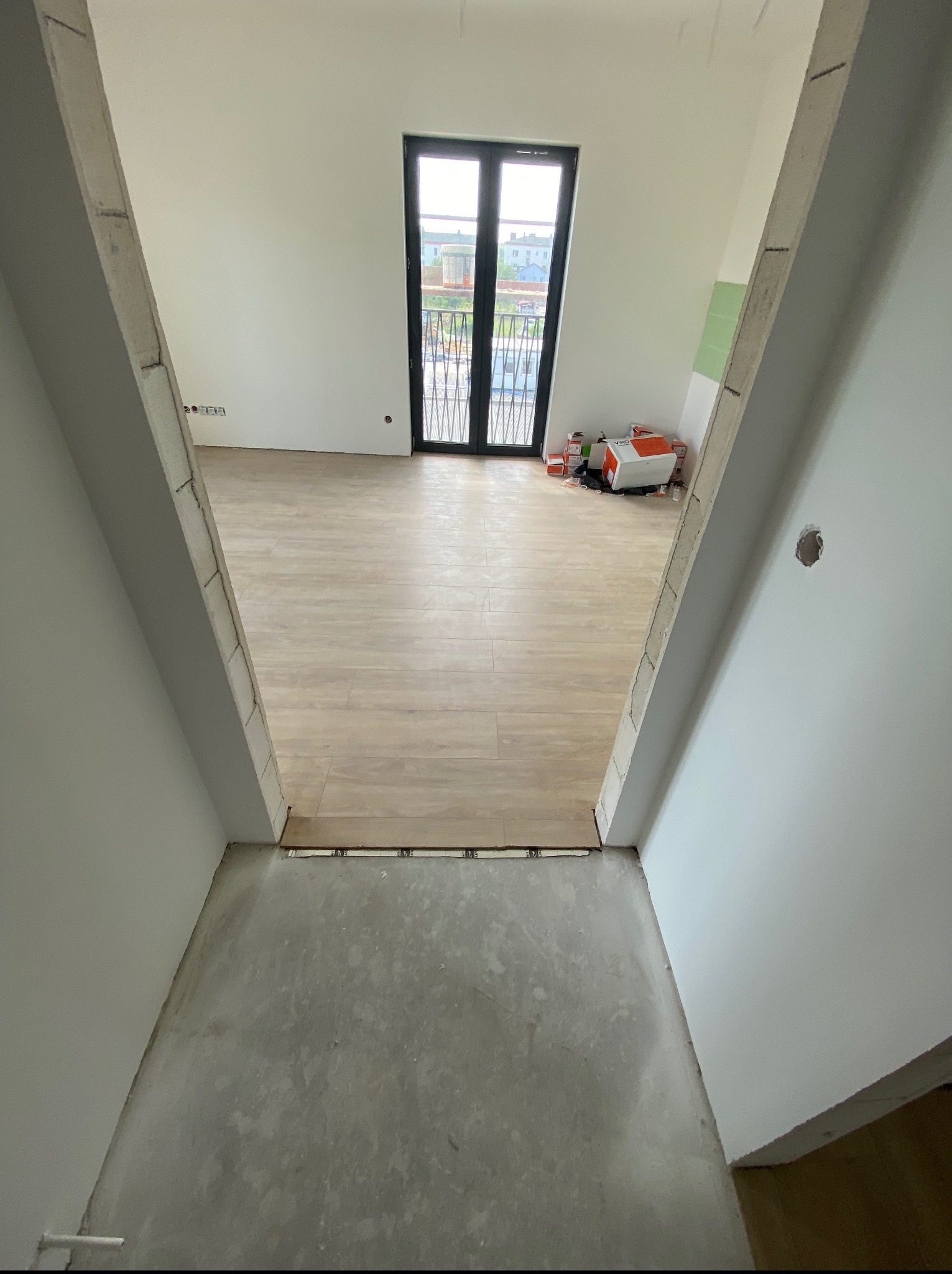 Wnętrze w trakcie remontu: ułożone panele podłogowe w pokoju z balkonem, surowy beton w przejściu, ściany przygotowane do malowania, widoczne puszki z farbą.