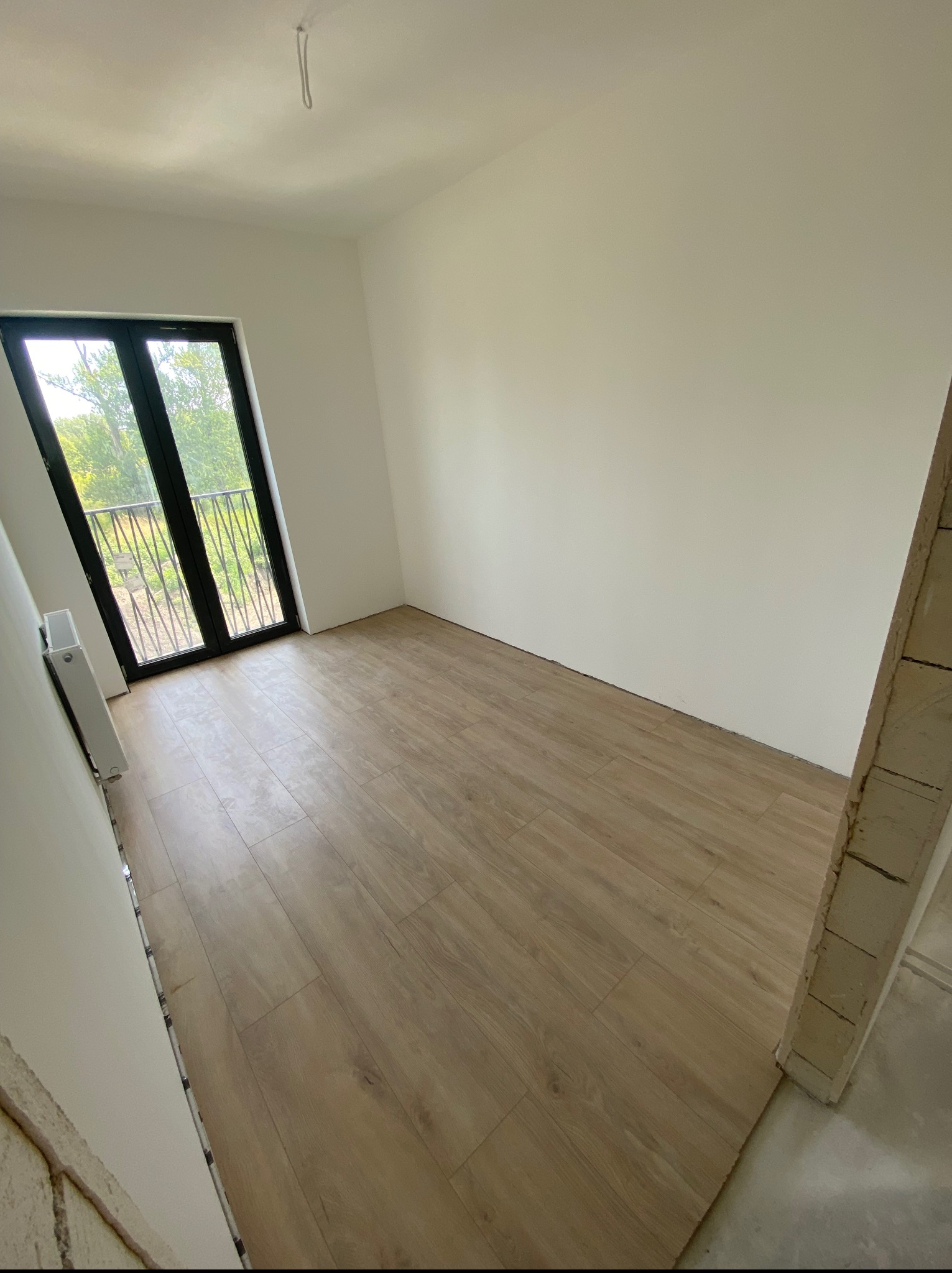 Jasne panele podłogowe w pustym pokoju z balkonem i białymi ścianami. Widoczny fragment surowej ściany. Minimalistyczne wnętrze w neutralnej tonacji.