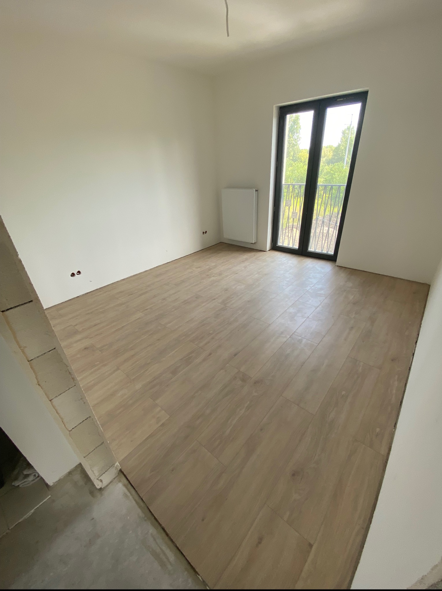 Jasne panele podłogowe w pokoju z balkonem i grzejnikiem. Widoczne gniazdka elektryczne i surowy fragment ściany. Naturalne światło dzienne.