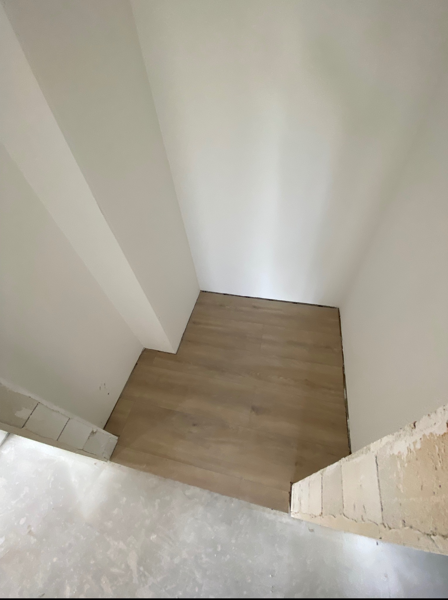 Fragment pokoju z ułożonymi panelami podłogowymi w odcieniach brązu, widok z góry, białe ściany i sufit, surowy stan wykończenia przy podłodze.