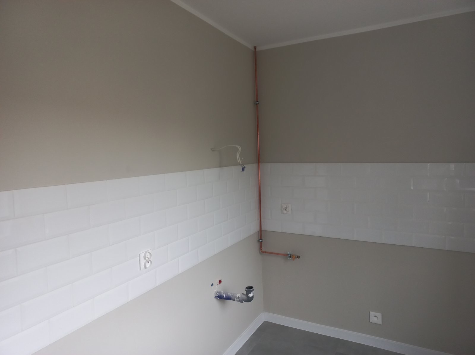 Ściana z białymi, prostokątnymi płytkami typu metro, szara ściana i miedziana instalacja wodna z przyłączem kanalizacyjnym w rogu pomieszczenia.