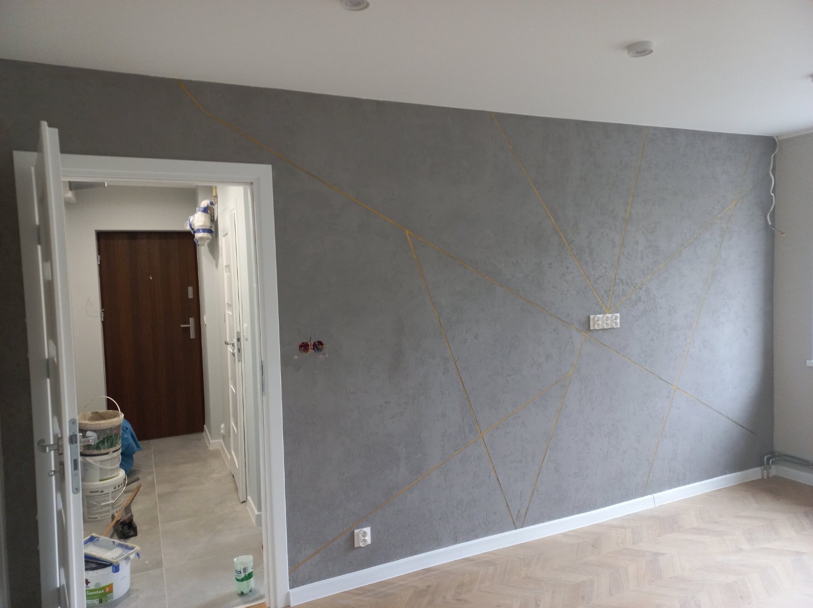Szara ściana z dekoracyjnym wzorem złotych linii, widok na wnętrze przez otwarte drzwi, parkiet na podłodze, puszki z farbą w tle.