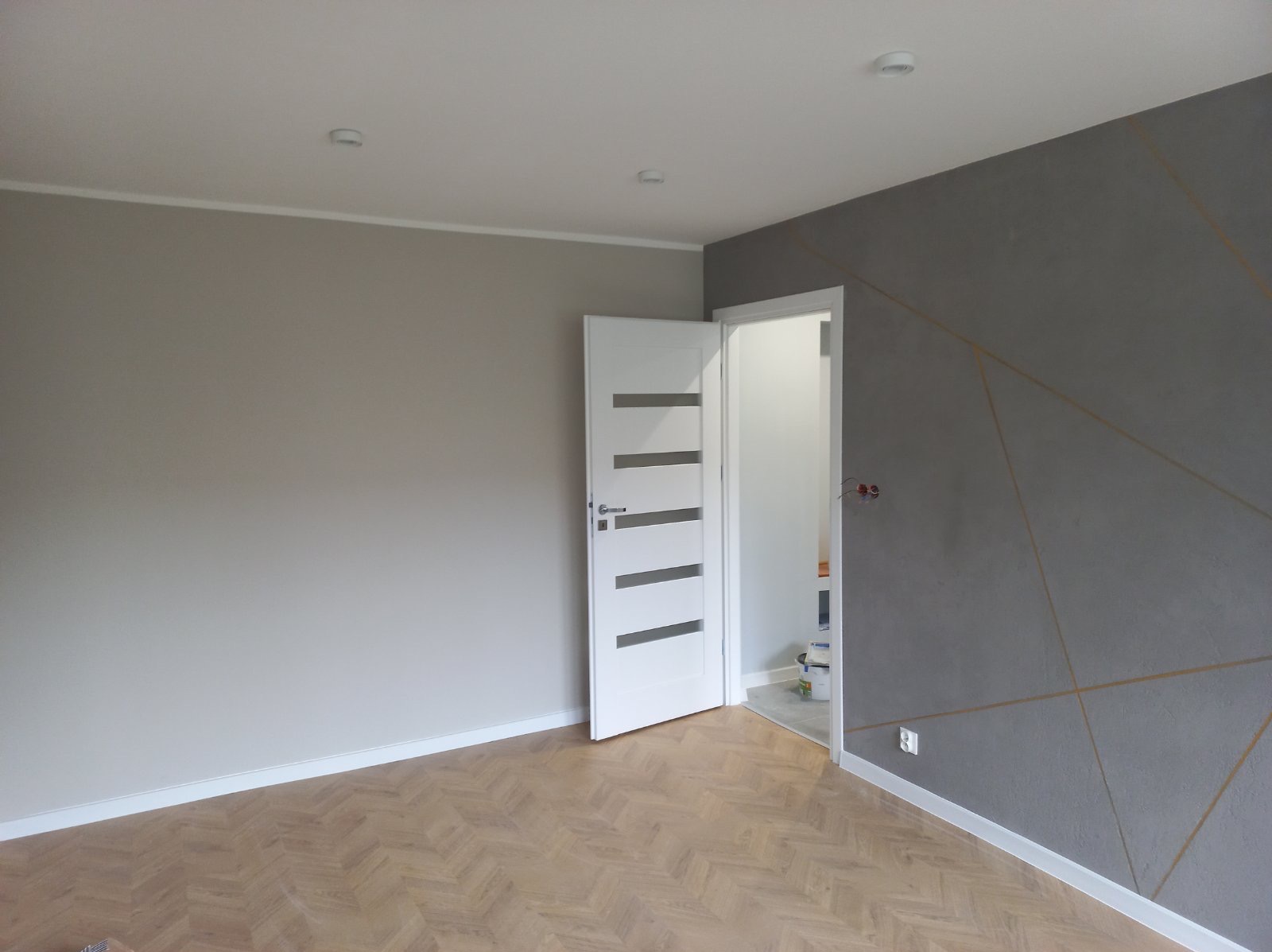 Pokój po remoncie: parkiet w jodełkę, szara ściana z geometrycznym wzorem, białe drzwi z poziomymi przeszkleniami. Widoczne detale wykończeniowe.