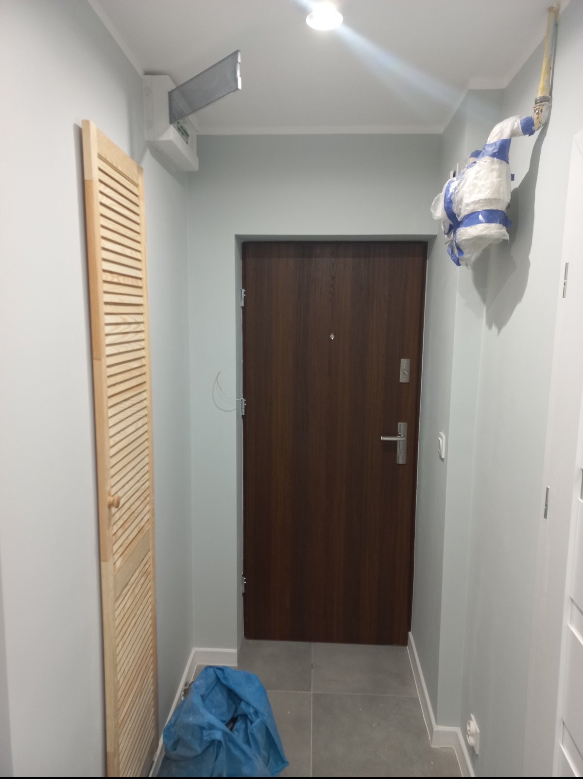 Korytarz po remoncie w Bytomiu: drewniana szafka z lamelami, brązowe drzwi wejściowe, szare ściany i podłoga z płytek. Widoczny klimatyzator i zabezpieczone rury.
