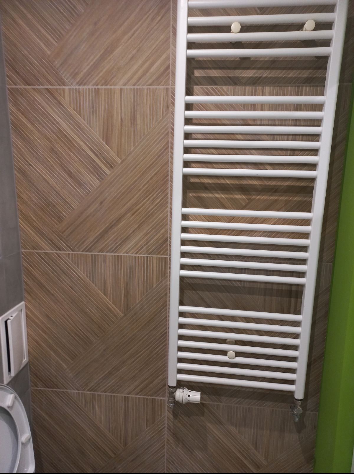 Biały grzejnik łazienkowy na tle ściany z płytek imitujących drewno ułożonych w geometryczny wzór. Widoczny fragment toalety i zielony element.