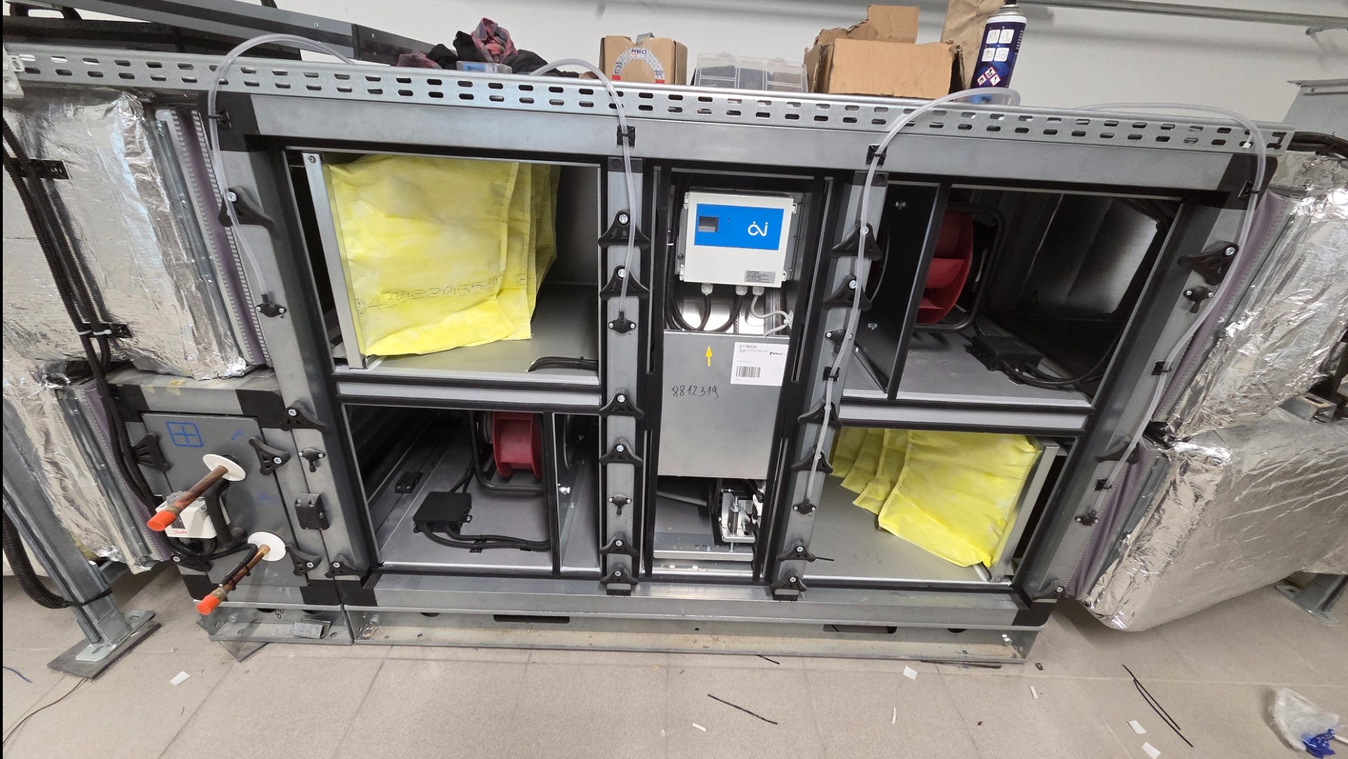 Centrala wentylacyjna w metalowej obudowie z widocznymi filtrami, wentylatorami i elementami sterującymi. Widoczne przewody i izolacja termiczna.