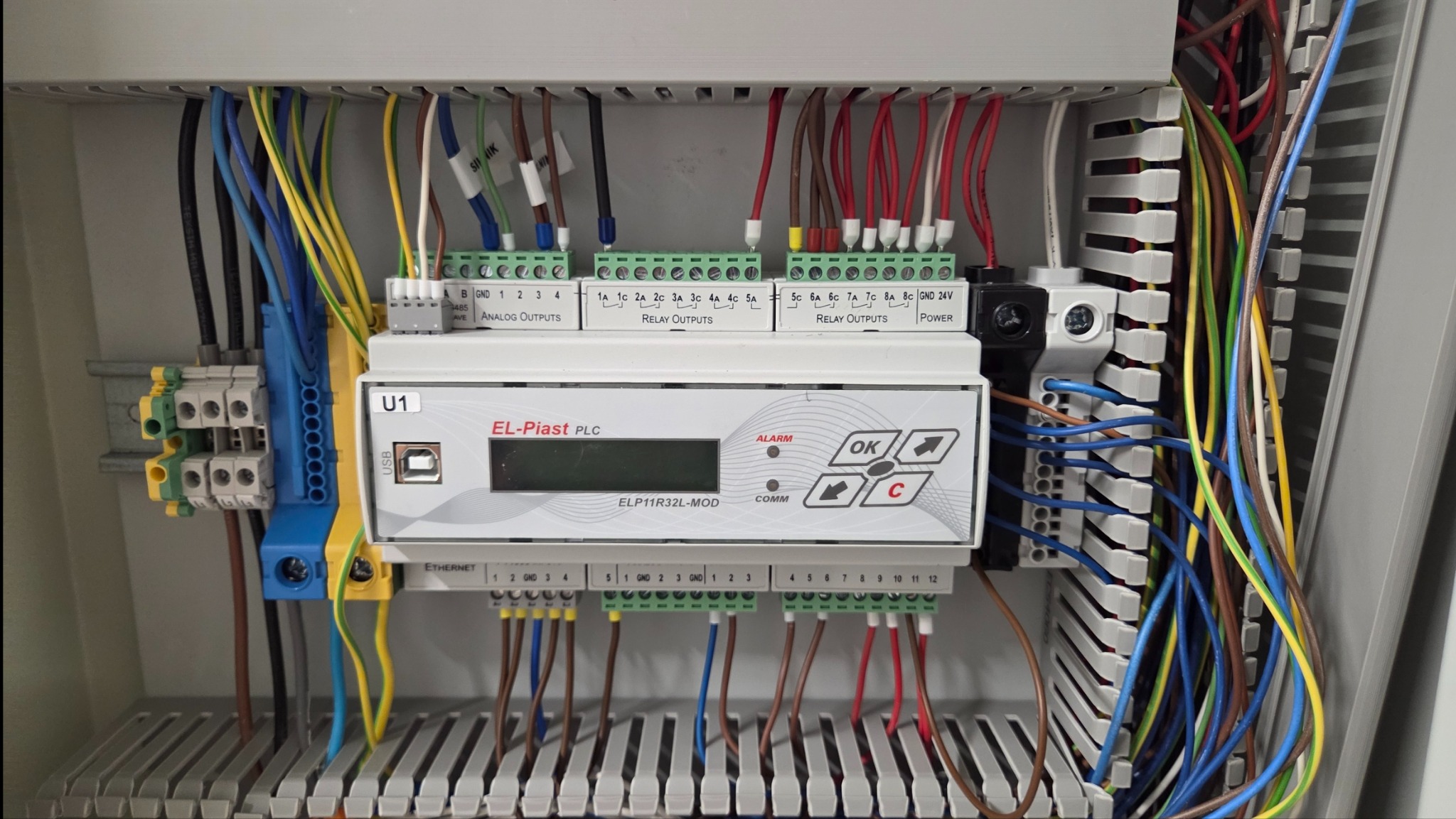 Panel sterowania PLC El-Piast z okablowaniem, widoczne złącza, przewody w różnych kolorach i oznaczenia. W tle szara obudowa.
