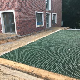 Daniel-Gmbh - Montaż ażurowych płyt trawnikowych na podsypce piaskowej przy nowym domu z czerwonej cegły. Widoczny fragment ułożonej kostki i narzędzie.