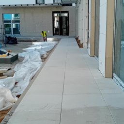 Daniel-Gmbh - Jasnoszara kostka brukowa ułożona wzdłuż budynku z pracownikiem w odblaskowej kamizelce w tle. Widoczne przygotowania do dalszych prac brukarskich.