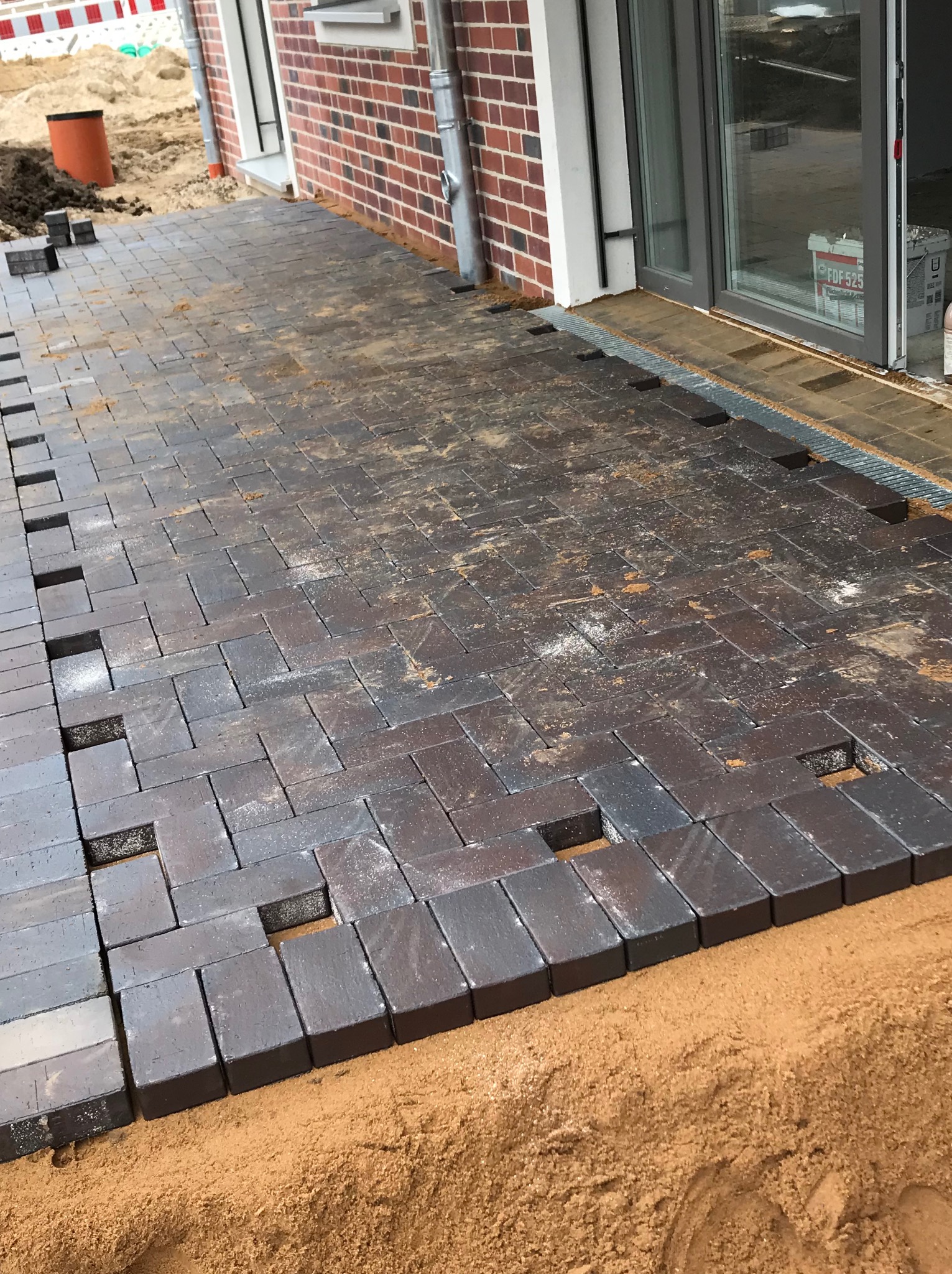 Ciemna kostka brukowa ułożona częściowo na piasku, z widocznymi przerwami między elementami. W tle fragment budynku z cegły i otwarte drzwi balkonowe.