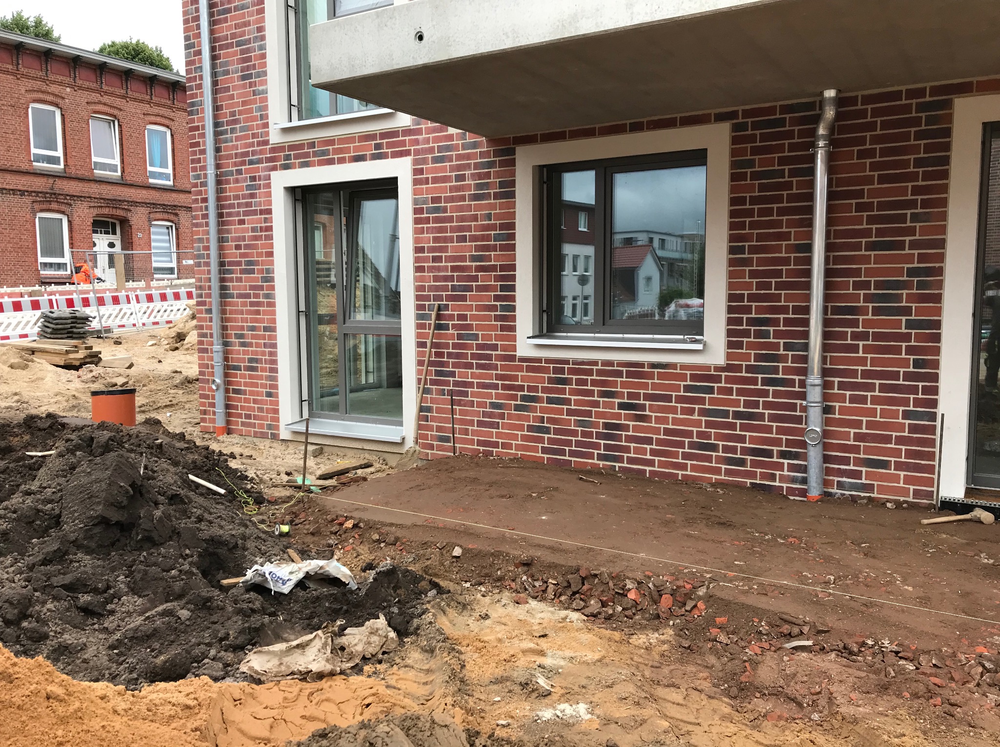 Przygotowanie terenu pod budowę: wykop, hałda ziemi, fragment muru z cegły. Widoczne narzędzia i elementy konstrukcyjne. Prace ziemne w toku.