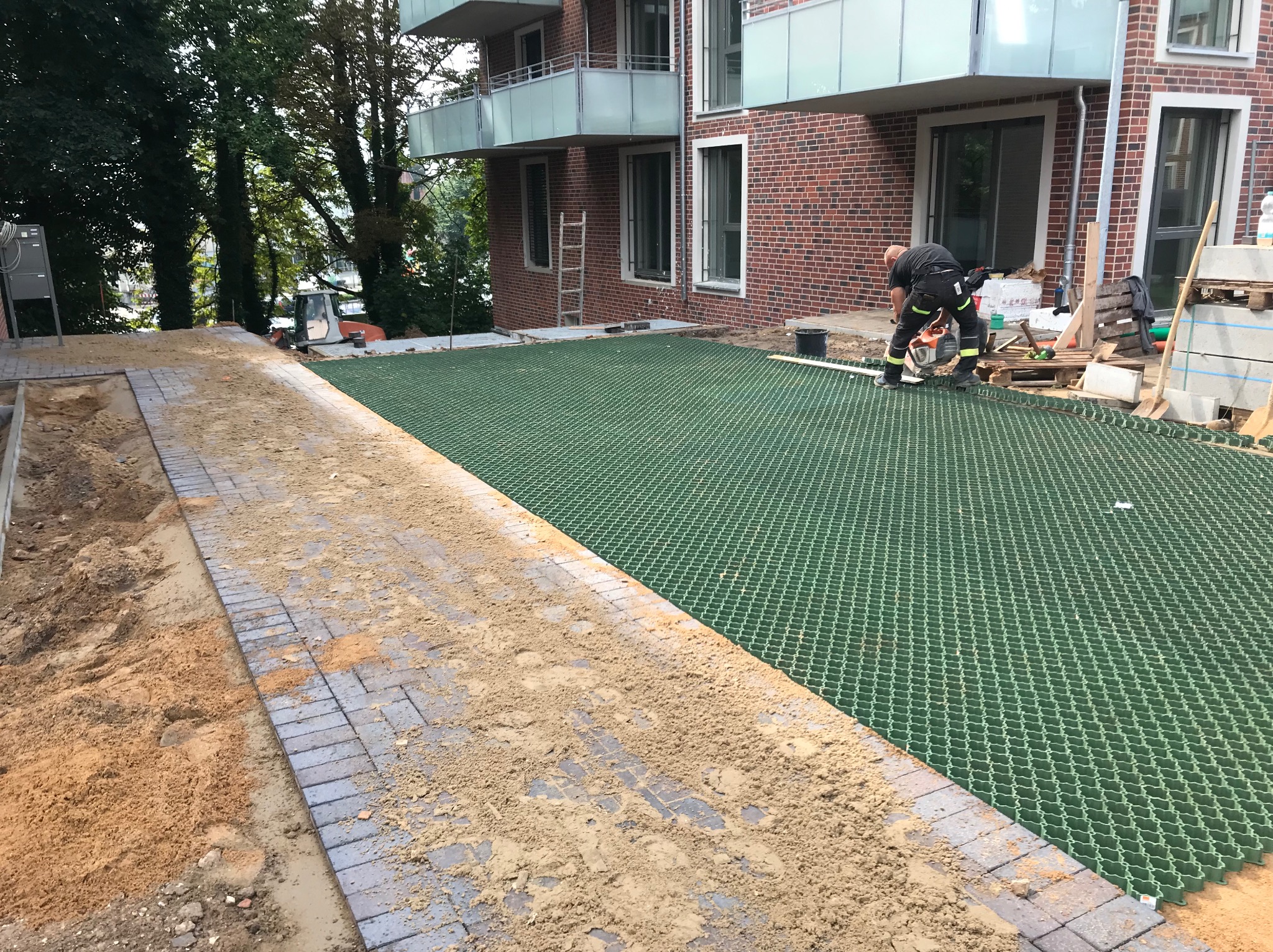 Montaż ażurowych płyt trawnikowych na podbudowie z kostki brukowej. Pracownik docina płytę piłą. Budynek w tle. Widoczne materiały i narzędzia.