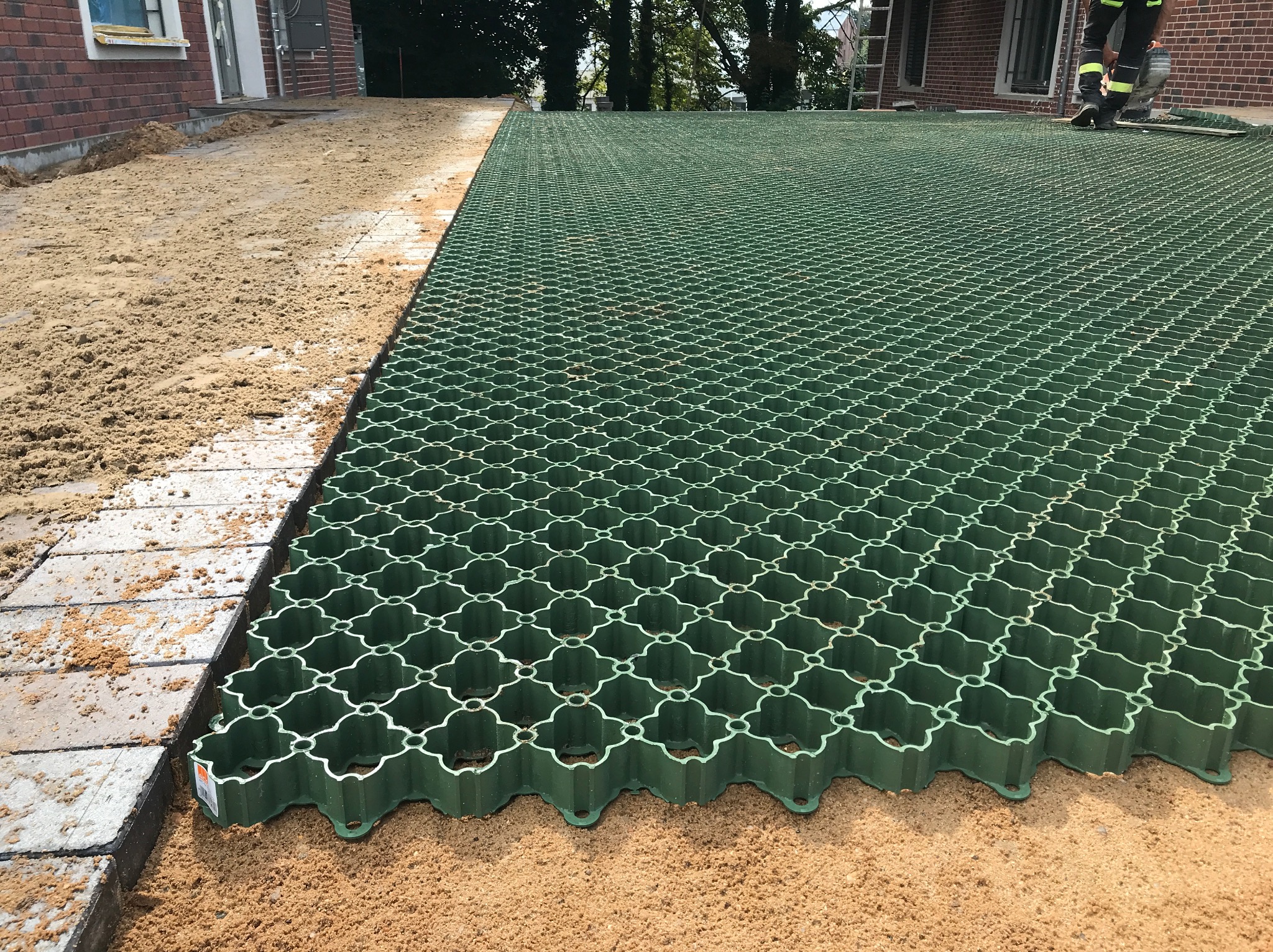 Montaż zielonej kratki trawnikowej na piasku, widoczne fragmenty kostki brukowej i budynku. Etap przygotowania podłoża, perspektywa z poziomu gruntu.