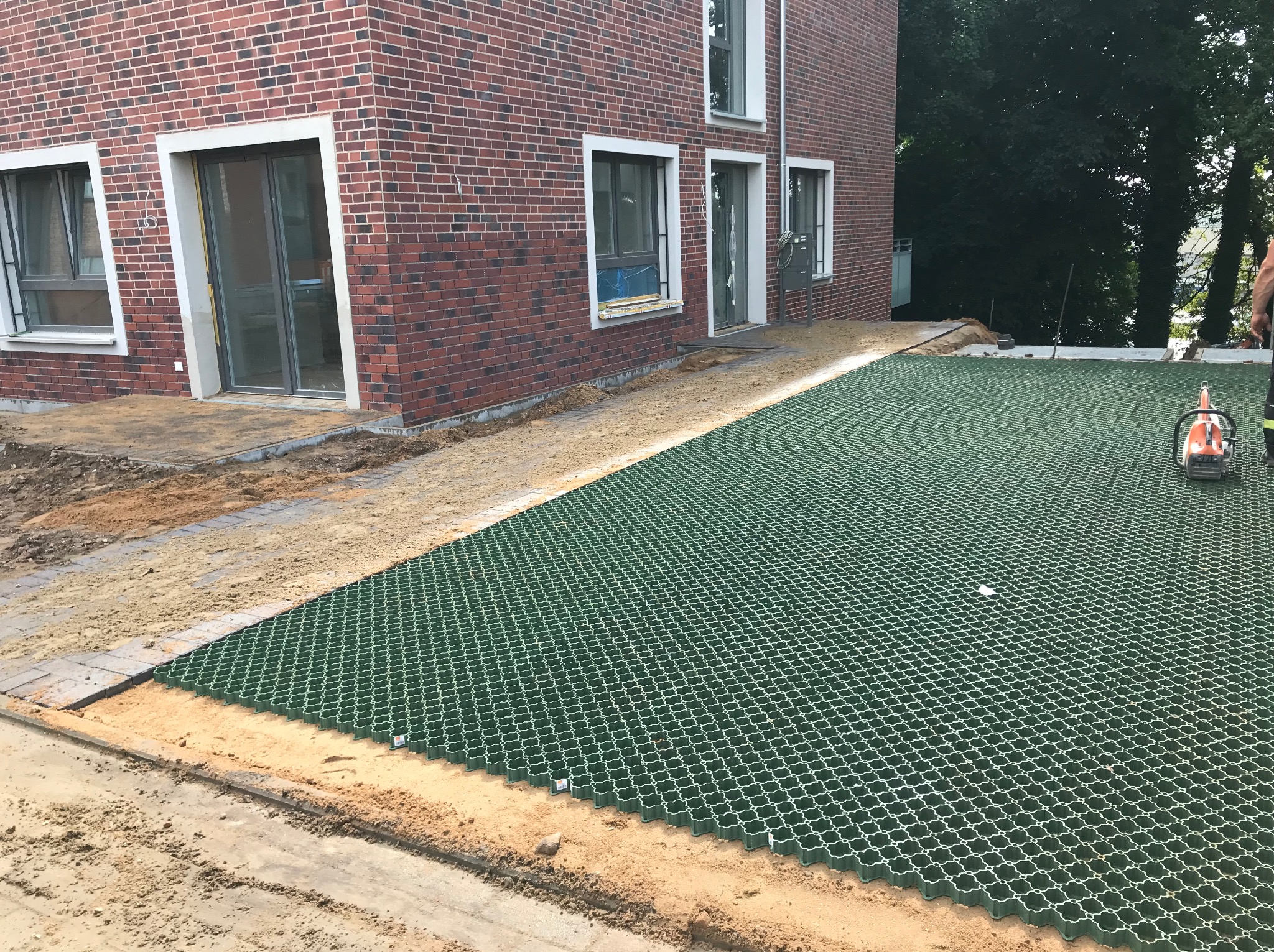 Montaż ażurowych płyt trawnikowych na podsypce piaskowej przy nowym domu z czerwonej cegły. Widoczny fragment ułożonej kostki i narzędzie.