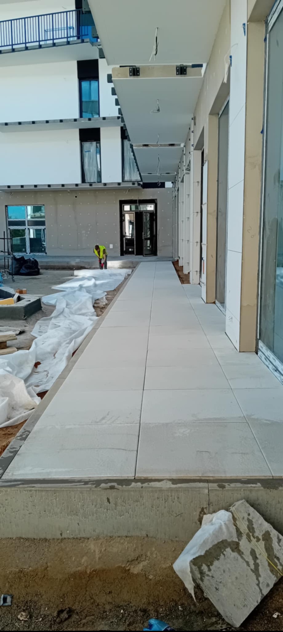 Jasnoszara kostka brukowa ułożona wzdłuż budynku z pracownikiem w odblaskowej kamizelce w tle. Widoczne przygotowania do dalszych prac brukarskich.