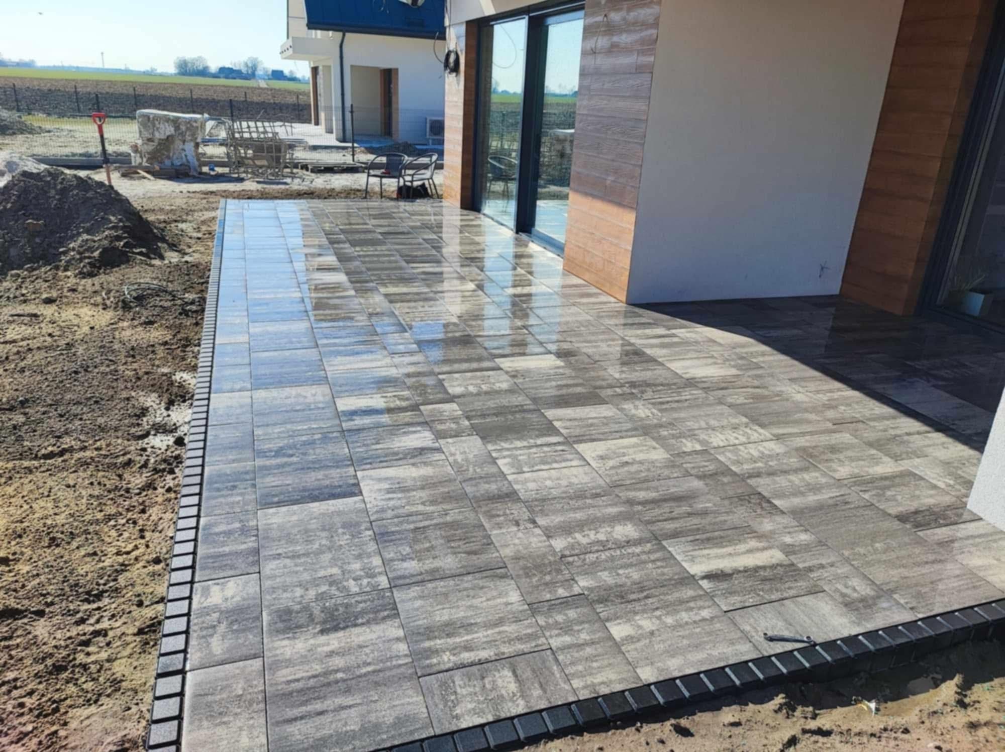 Nowoczesny taras z szarej kostki brukowej z dekoracyjnym obrzeżem, przylegający do domu z dużymi oknami przesuwnymi i elementami drewnianej elewacji.