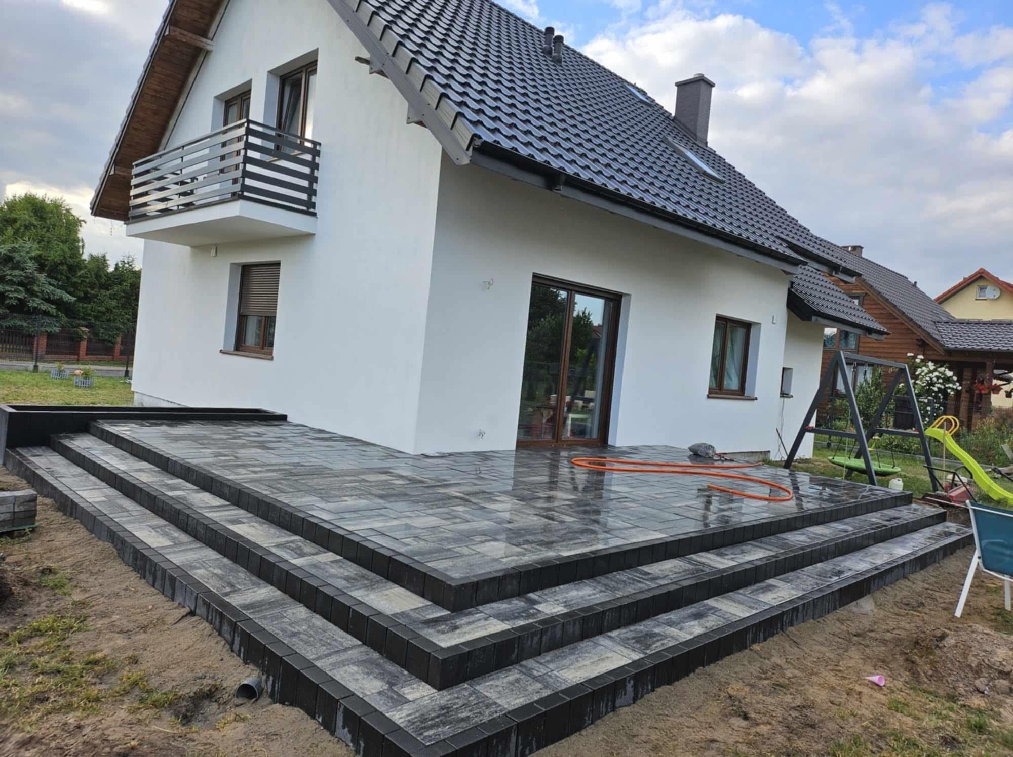 Nowoczesny taras z kostki brukowej w odcieniach szarości, prowadzący do białego domu z balkonem. Widoczny fragment ogrodu i błękitne niebo.
