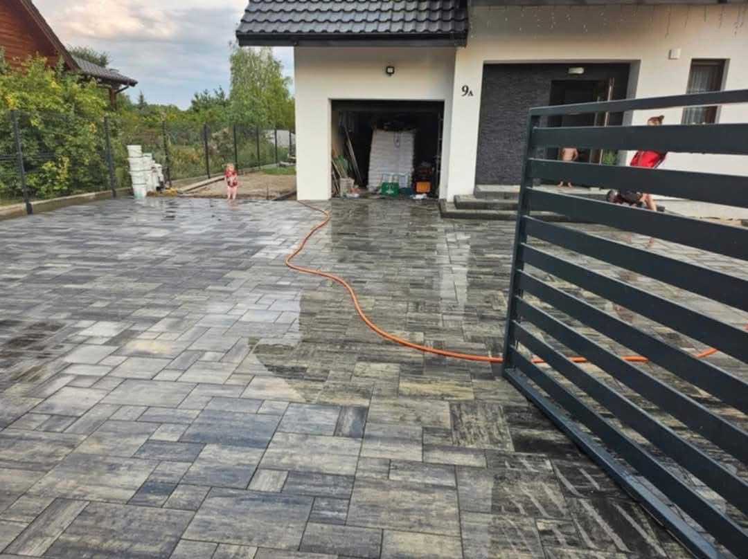 Ułożona kostka brukowa na podjeździe domu w szaro-grafitowej tonacji, z widocznym fragmentem ogrodzenia i wężem ogrodowym. W tle dom z otwartym garażem.