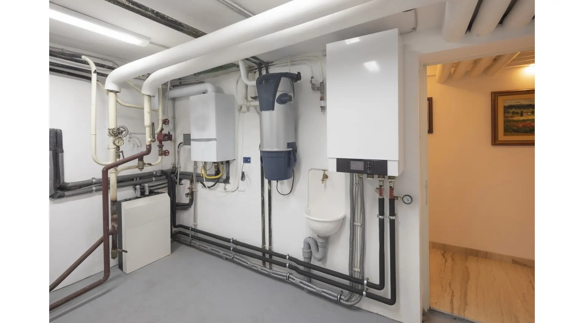 Piwnica z zainstalowanymi piecami gazowymi, rurami grzewczymi i małą umywalką. Widoczne elementy instalacji CO oraz fragment korytarza z obrazem na ścianie.