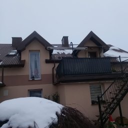 Marcin Głodowski - Dwukondygnacyjny dom z brązowym dachem z blachy trapezowej i oknami dachowymi, widoczny w zimowej scenerii. Na dachu leży śnieg.