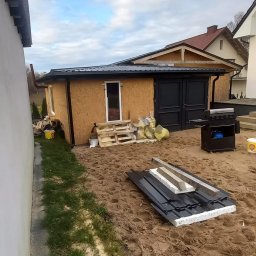 Marcin Głodowski - Budynek gospodarczy z nowym, czarnym dachem z blachy trapezowej. Na ziemi leżą przygotowane arkusze blachy. Widoczne rynny i obróbki blacharskie.