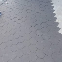 Marcin Głodowski - Układanie czarnej dachówki bitumicznej na płaskim dachu. Widoczne narzędzia i fragment buta dekarza. Częściowo pokryty dach z widocznymi rzędami dachówek.