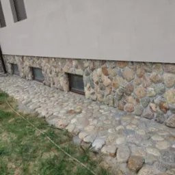 Wykończenie elewacji kamieniem rzecznym i ułożona z niego ścieżka wokół domu. Widoczne okna piwniczne i fragment trawnika. Kamień w różnych odcieniach szarości i beżu.