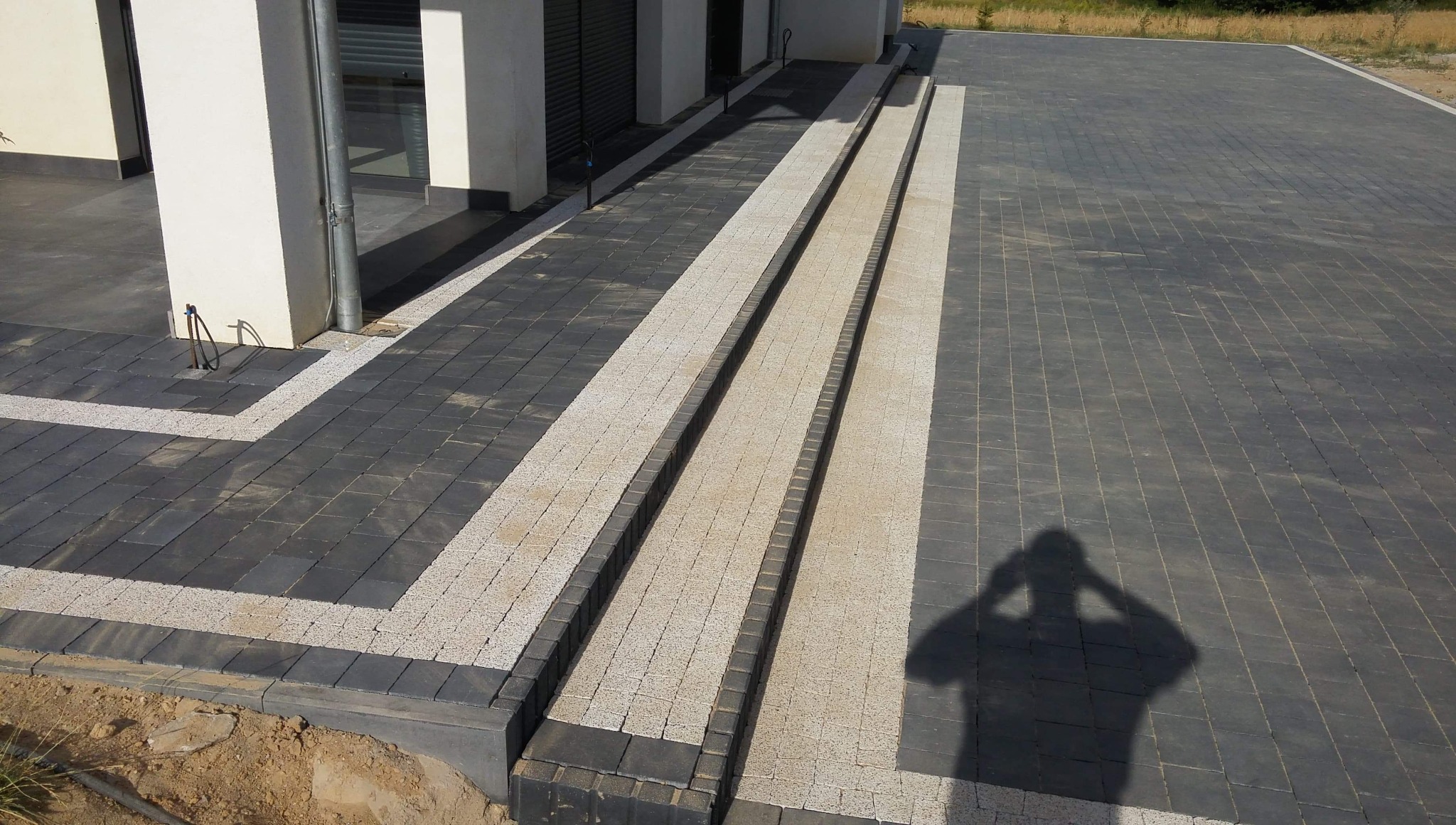 Nowoczesna nawierzchnia z kostki brukowej w odcieniach szarości i beżu, ułożona przed budynkiem. Widoczny cień osoby fotografującej.
